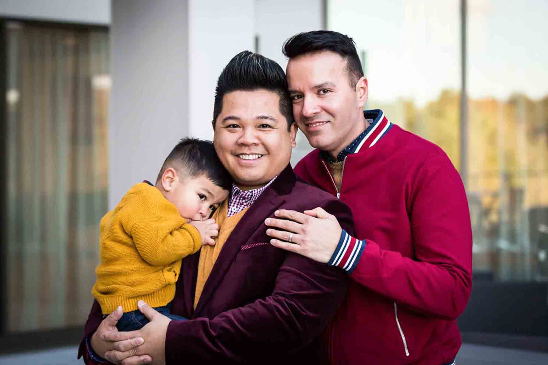 Family of two gay fathers holding little boy sucking his thumb in front of window for an article on daddy and me photo shoot ideas