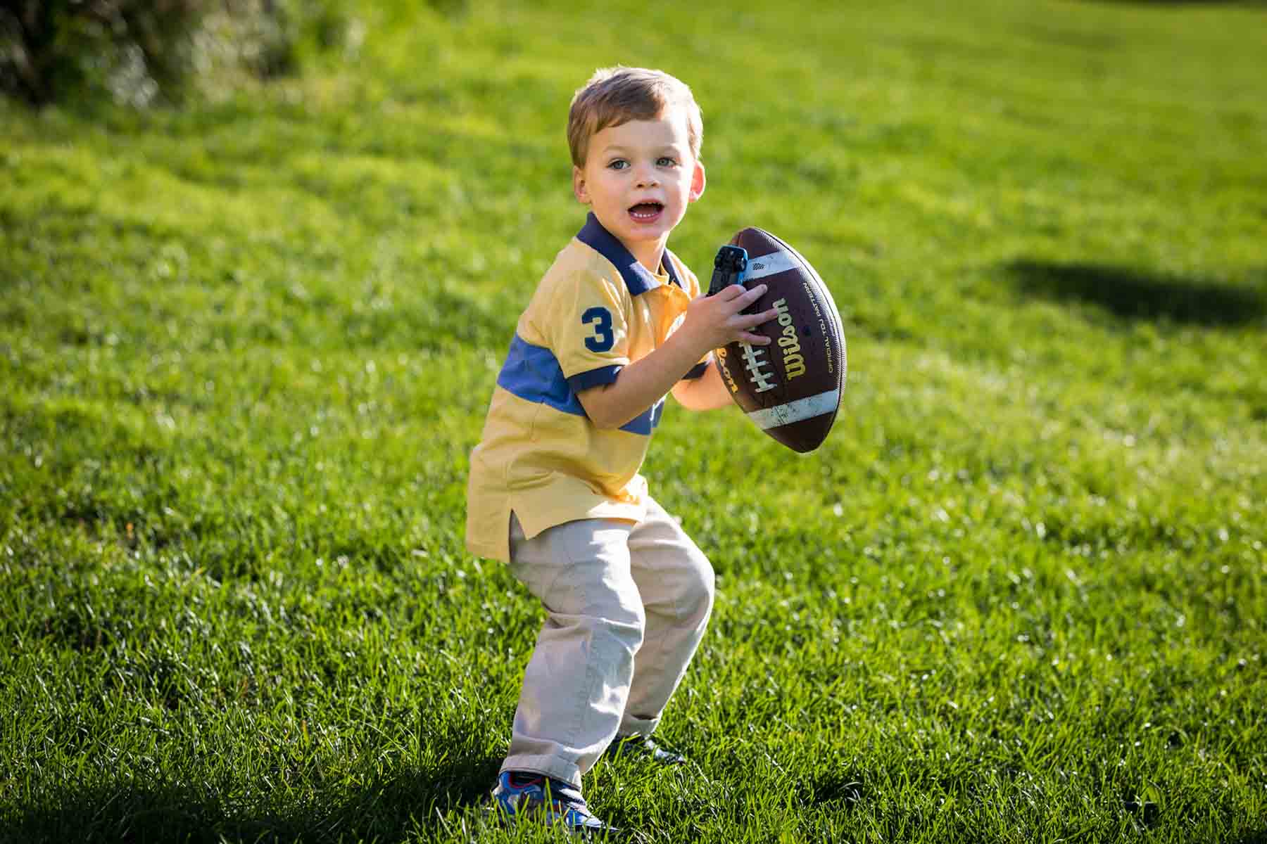 Little boy wearing yellow and blue shirt and holding football and toy car in field of grass for an article on daddy and me photo shoot ideas