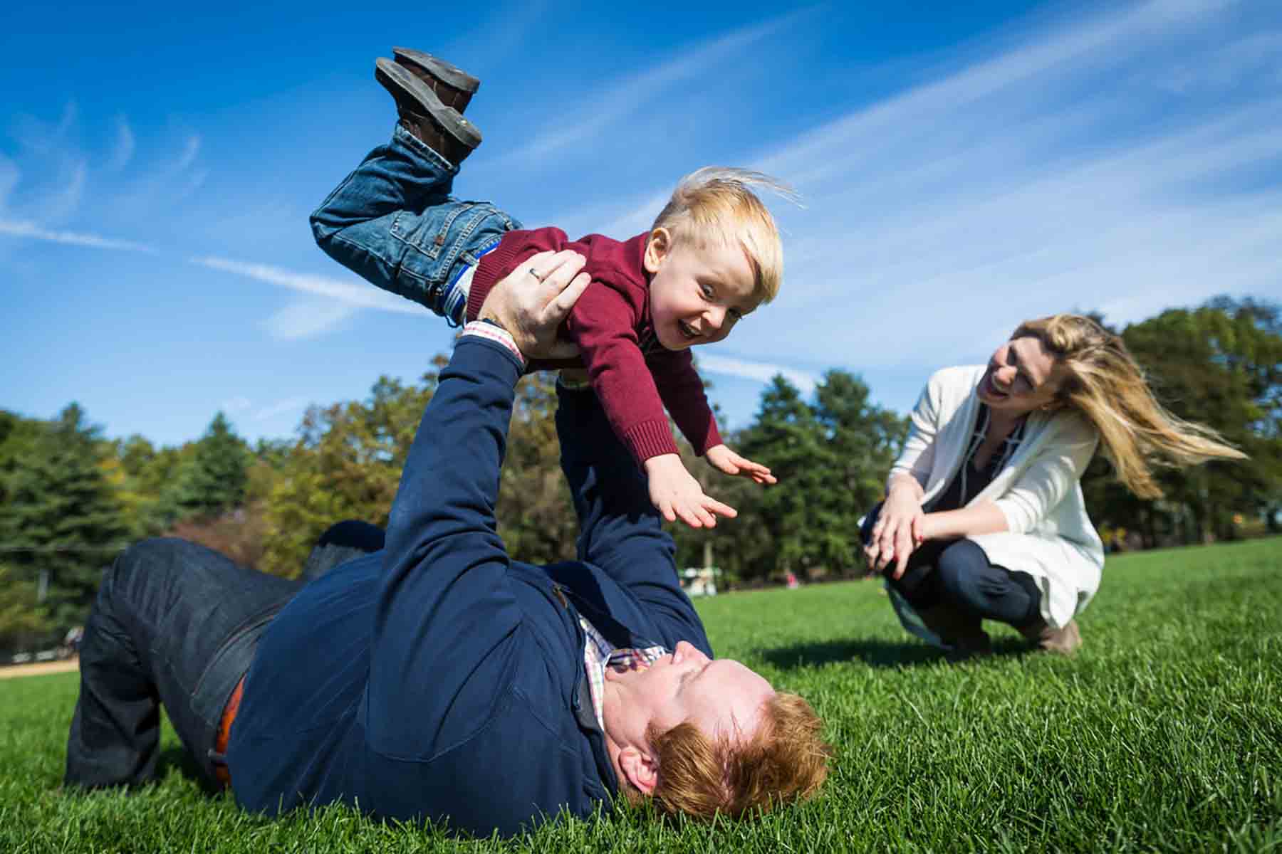 Father laying in grass holding up son in the air with mother watching to the right for an article on daddy and me photo shoot ideas