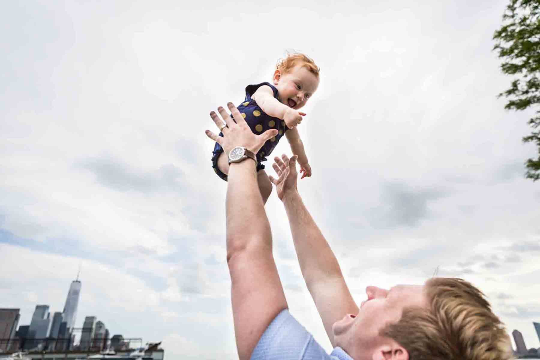 Father throwing little baby with red hair into the air with NYC skyline in the background for an article on daddy and me photo shoot ideas