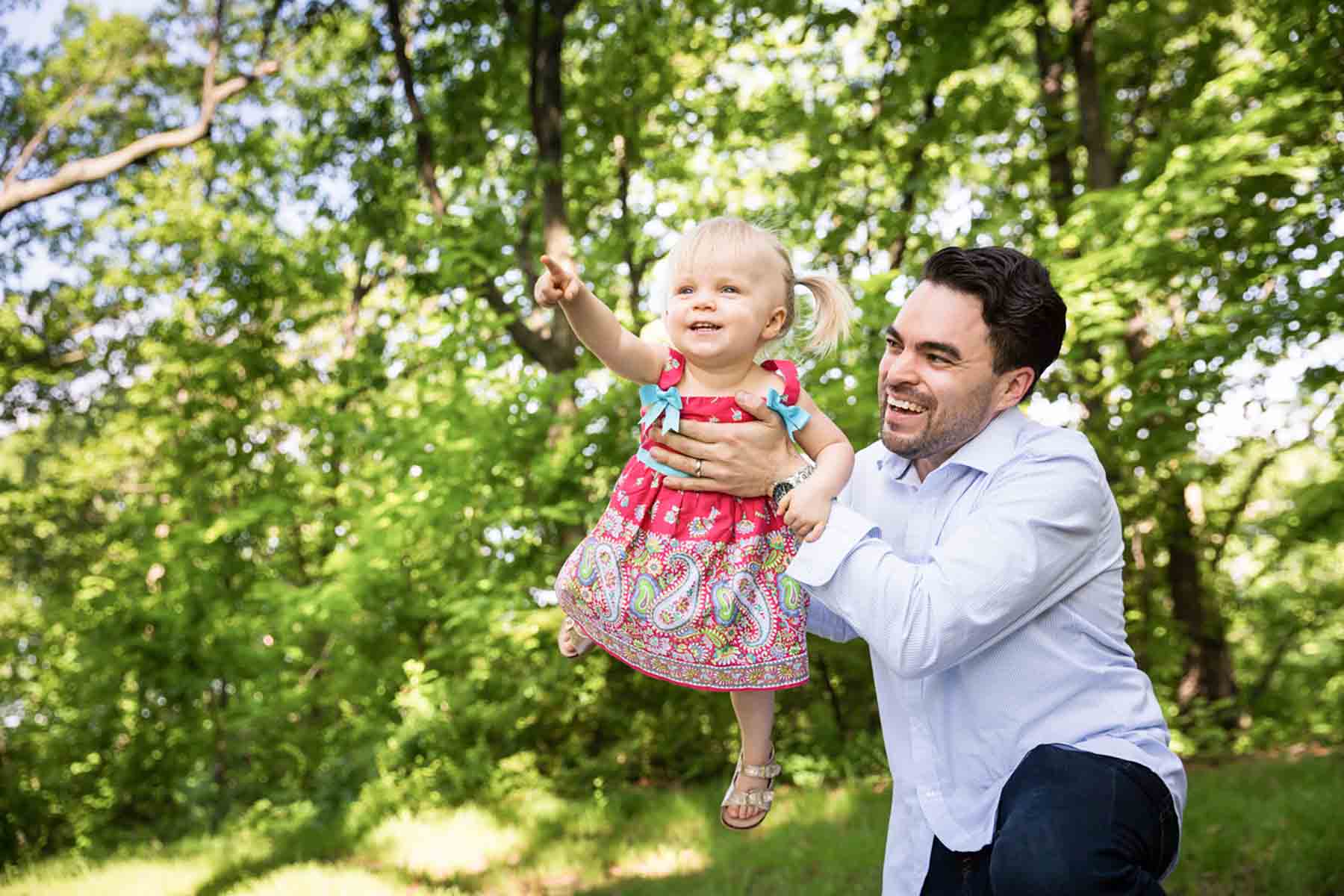 Father holding little blonde girl wearing pigtails and pointing in front of trees for an article on daddy and me photo shoot ideas