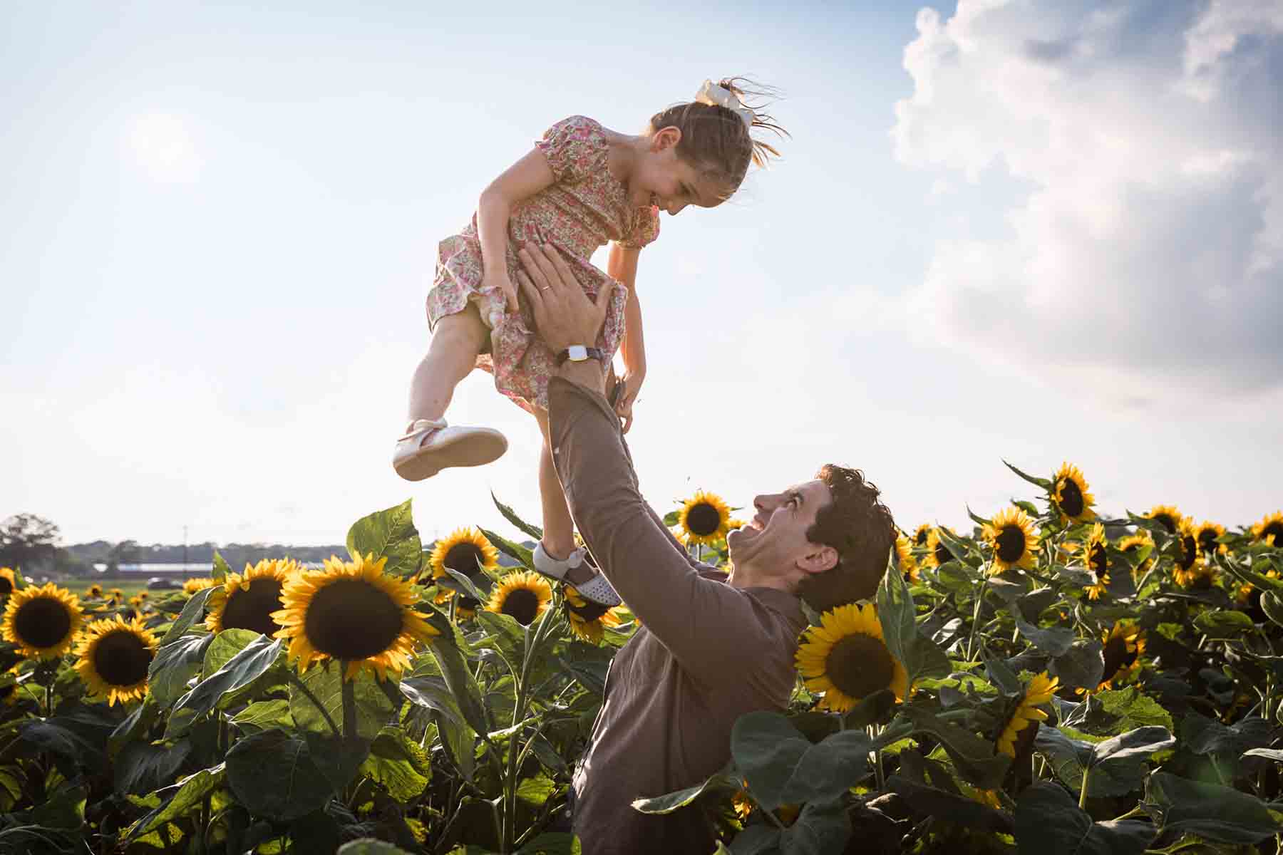 Father throwing little girl in the air in garden of sunflowers for an article on daddy and me photo shoot ideas