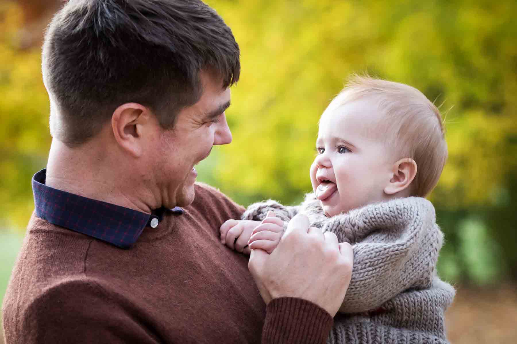 Little girl wearing beige sweater showing tongue to father in front of yellow bushes for an article on daddy and me photo shoot ideas