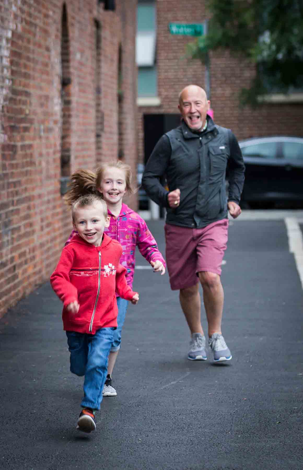 Bald man in shorts chasing after little boy and girl in front of brick wall for an article on daddy and me photo shoot ideas