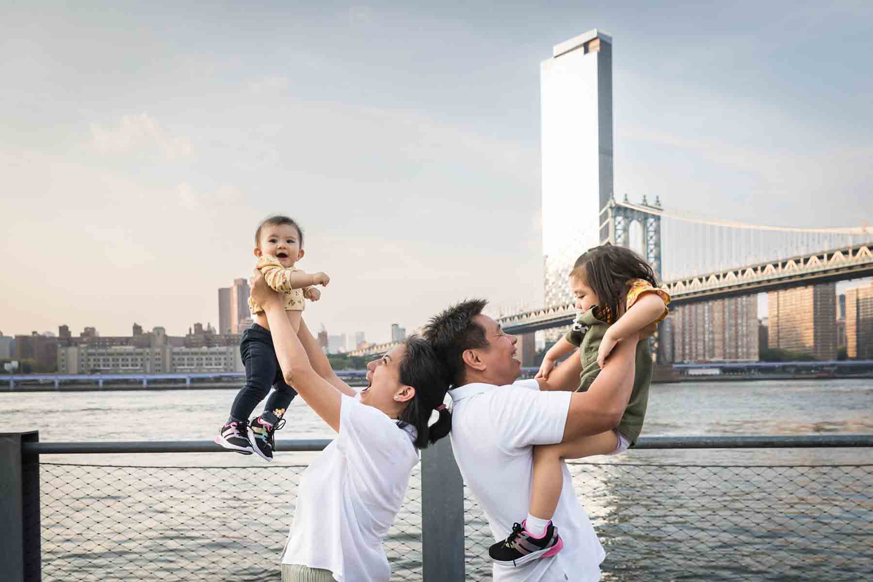 Mother and father holding up two little children with NYC skyline in the background for an article on daddy and me photo shoot ideas