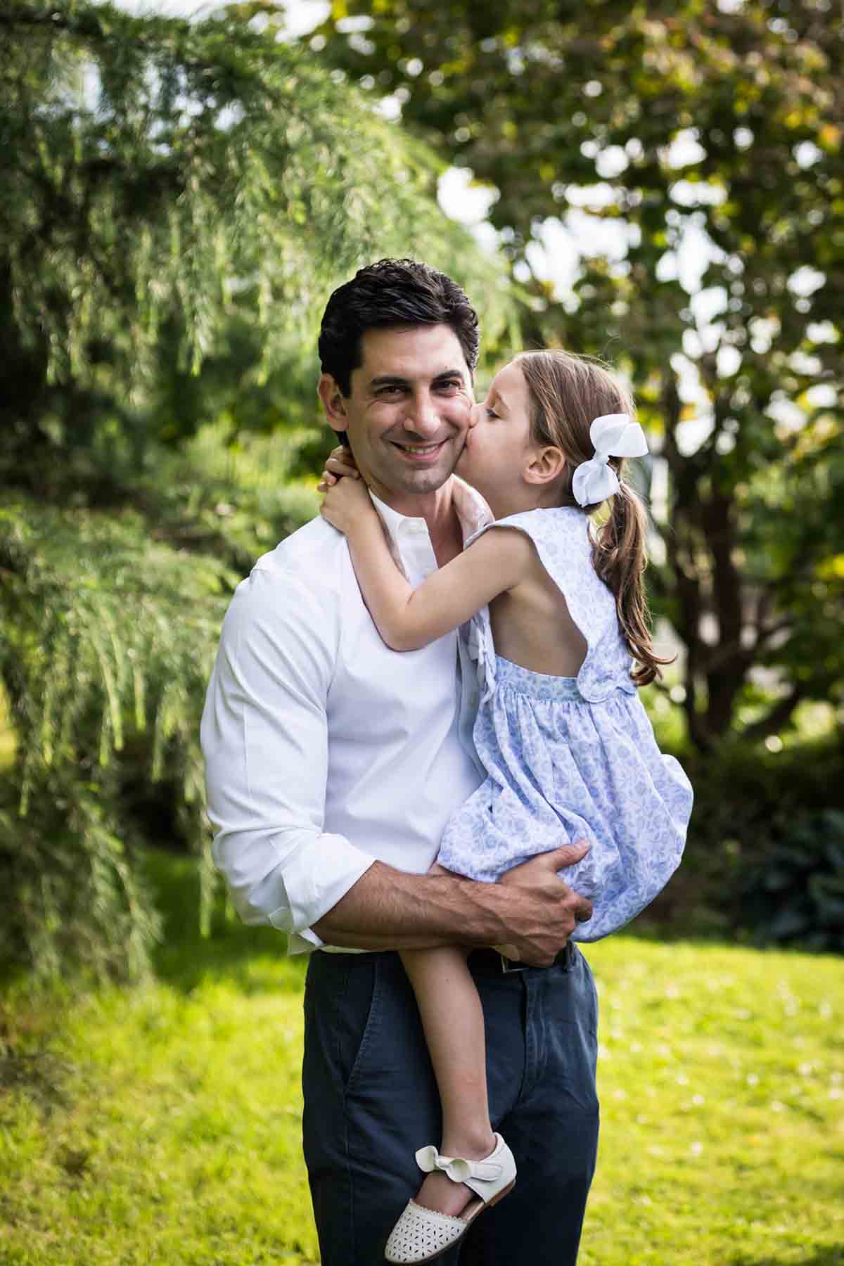 Father holding little girl and she is kissing him on the cheek in a park for an article on daddy and me photo shoot ideas