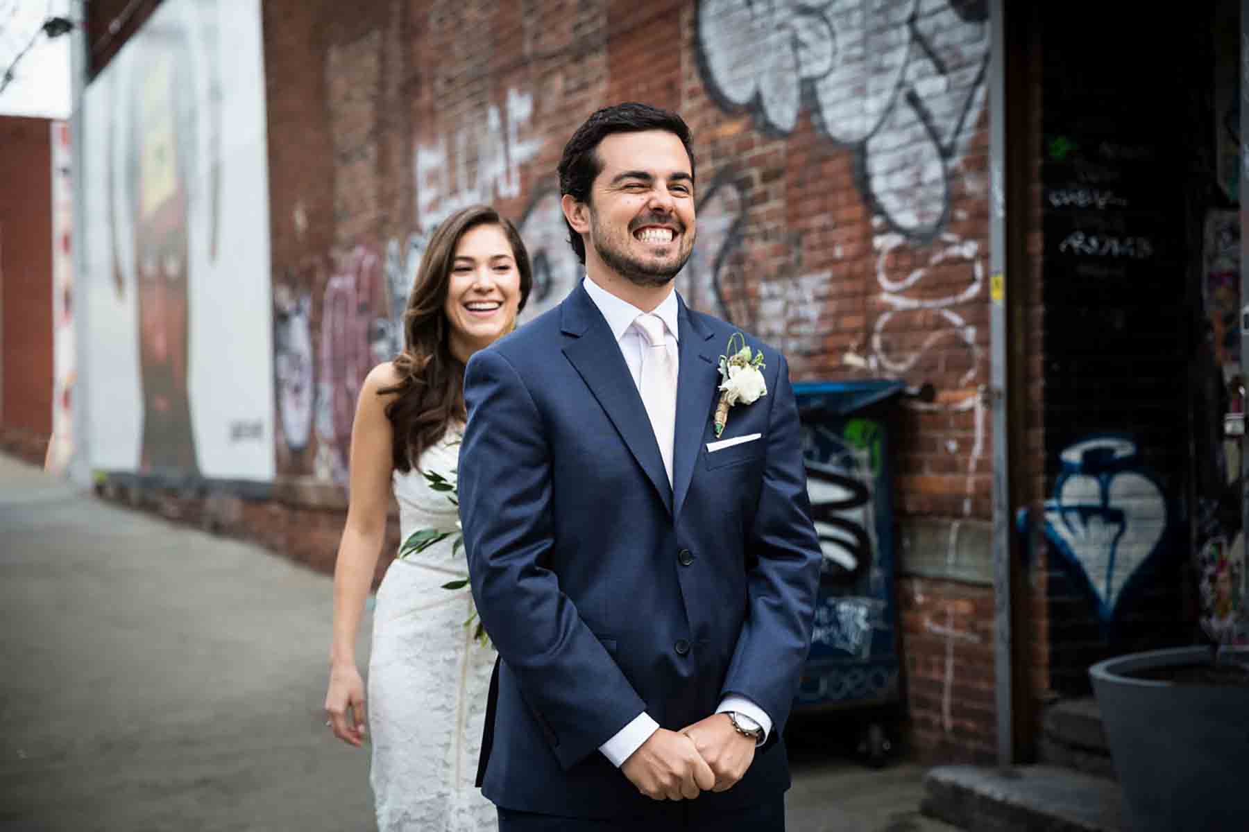 Bride sneaking up behind groom on sidewalk during first look for an article on how to save money on wedding photography
