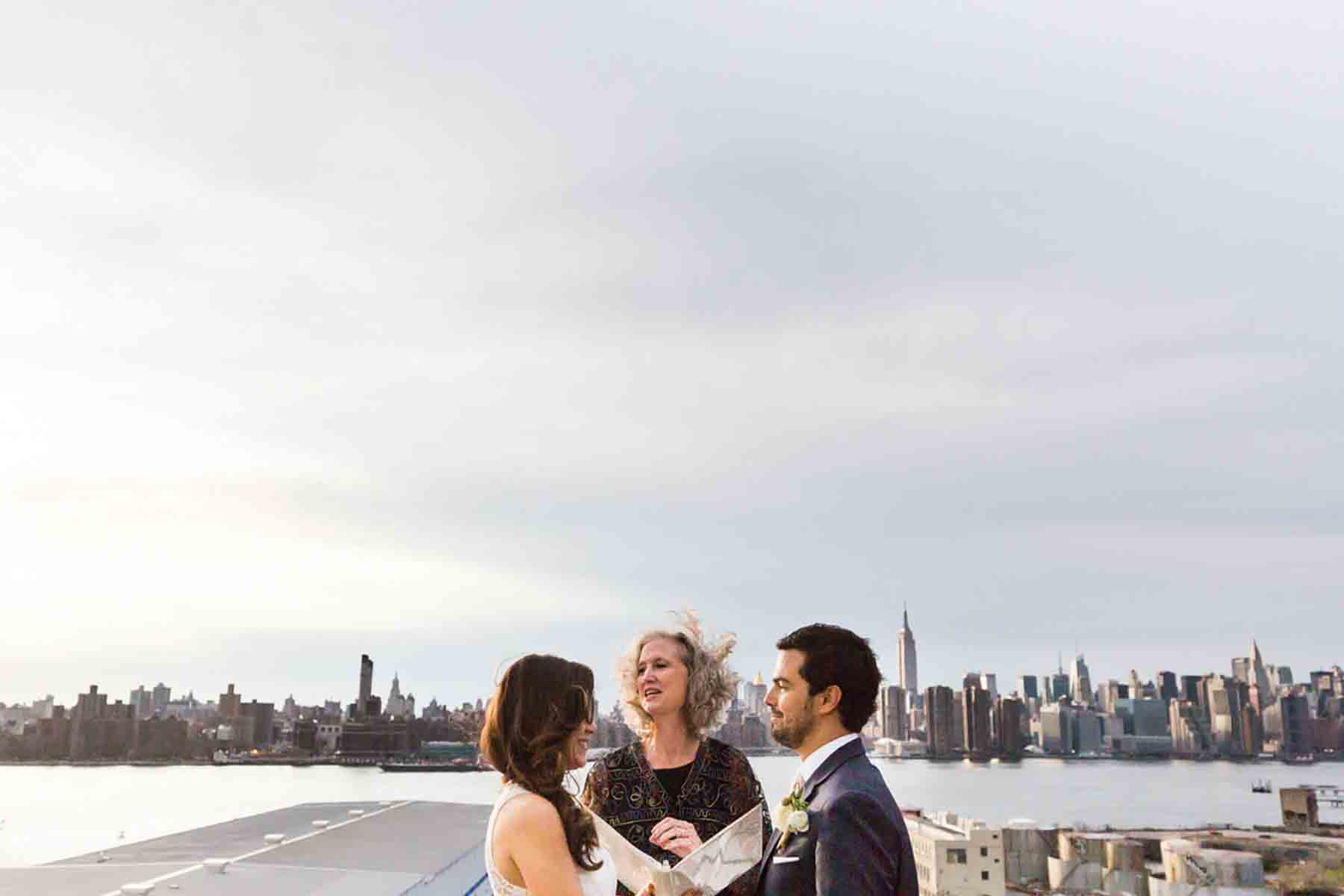 Bride and groom standing in front of officiant with NYC skyline in background for an article on how to save money on wedding photography