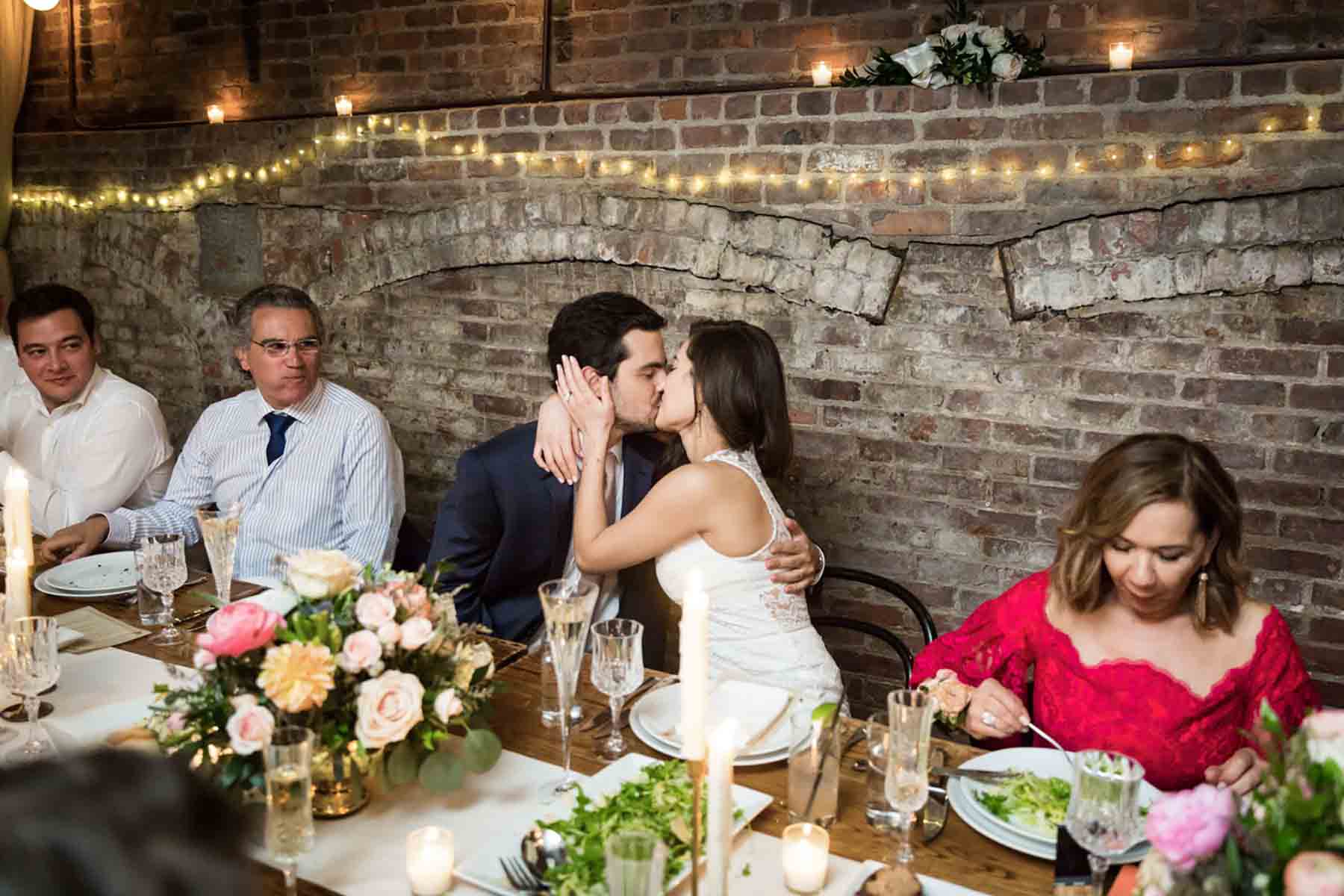 Bride and groom kissing at table in front of brick wall for an article on how to save money on wedding photography