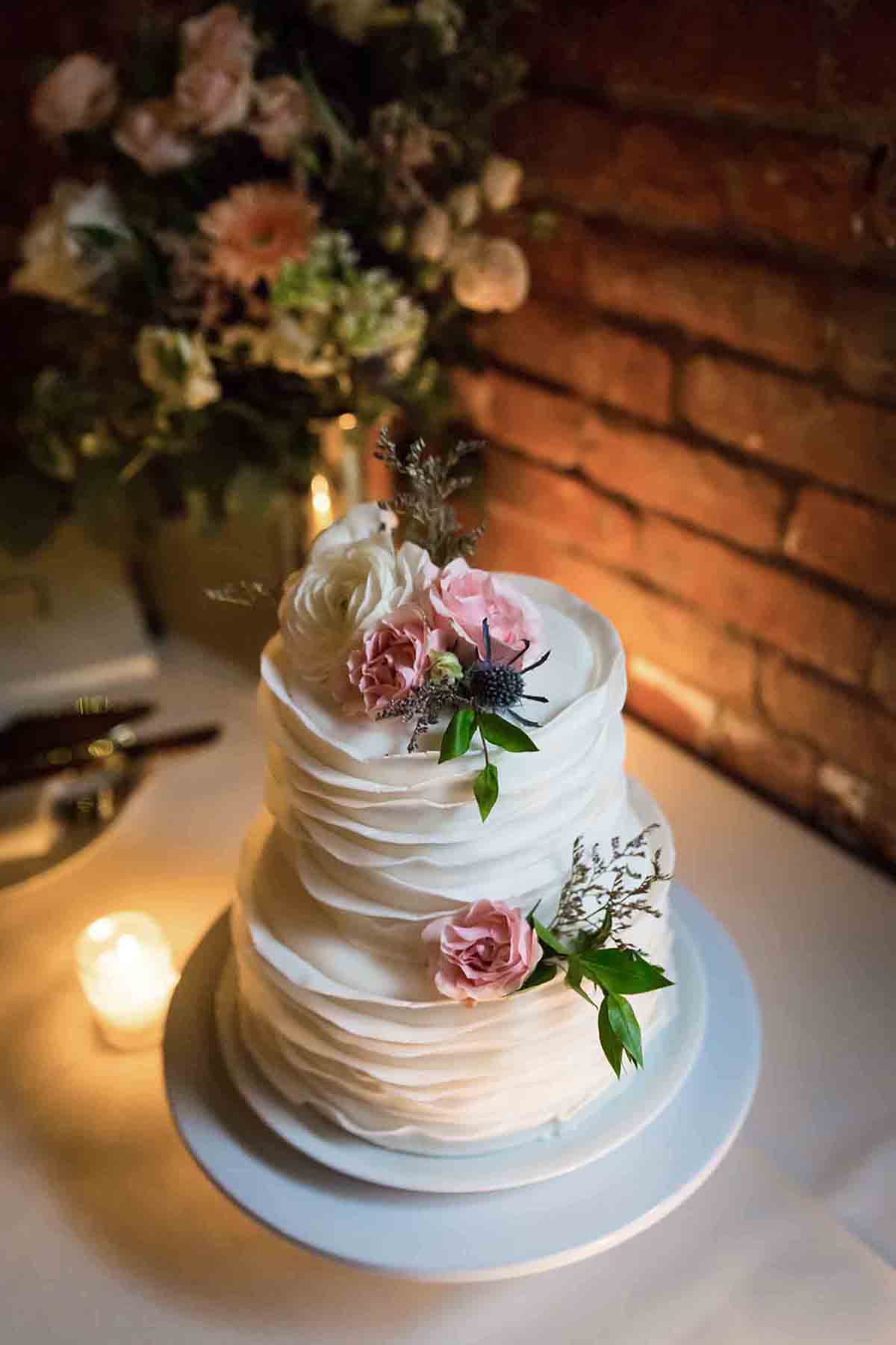 Two-tiered white wedding cake with flowers in front of brick wall for an article on how to save money on wedding photography