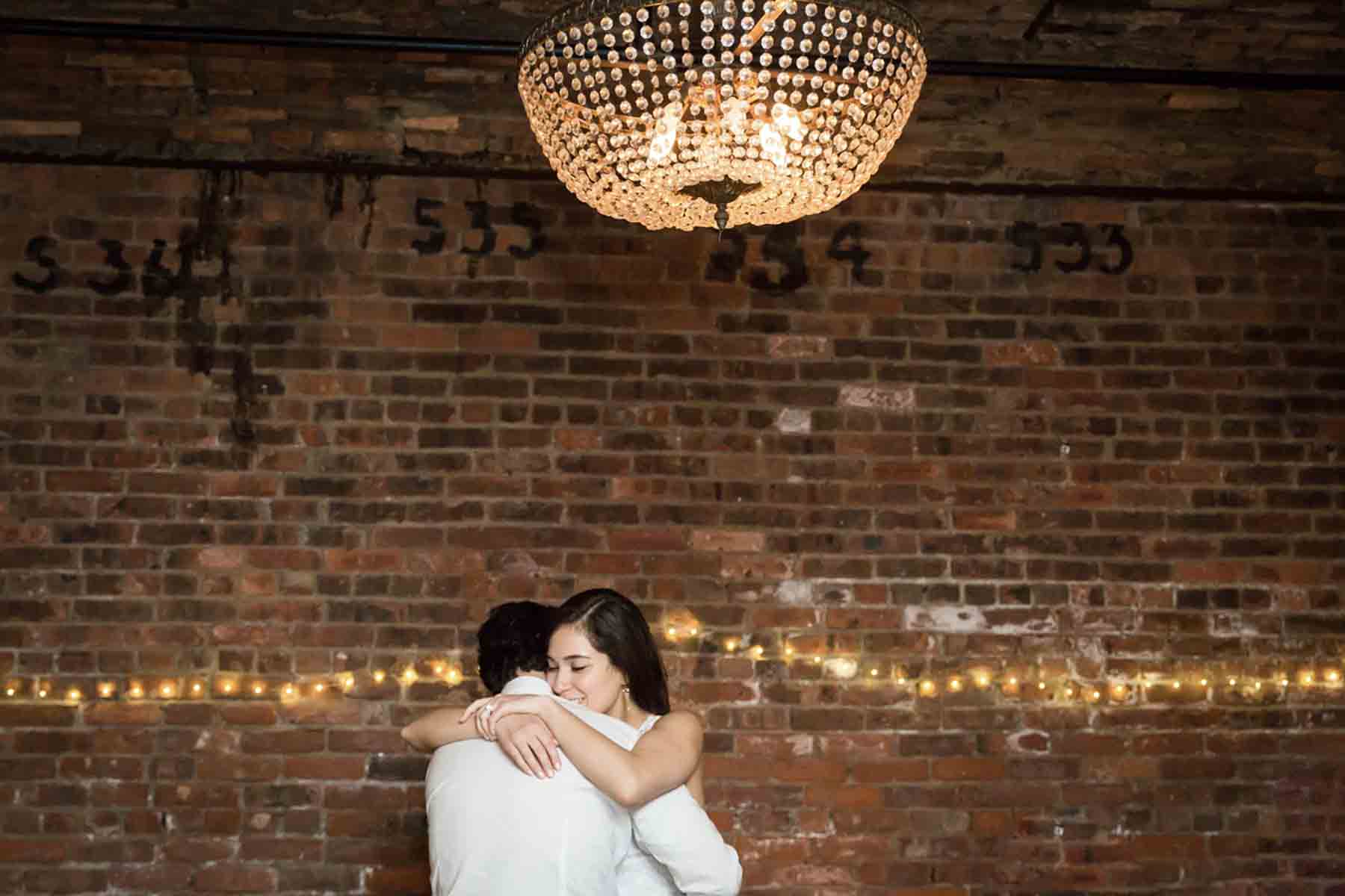 Bride and groom dancing under chandelier in front of brick wall for an article on how to save money on wedding photography