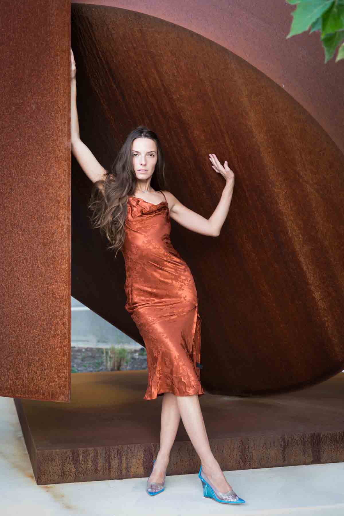 Model in rust-colored dress posing in front of rusted sculpture for an article announcing a downtown San Antonio photo shoot neighborhood discount