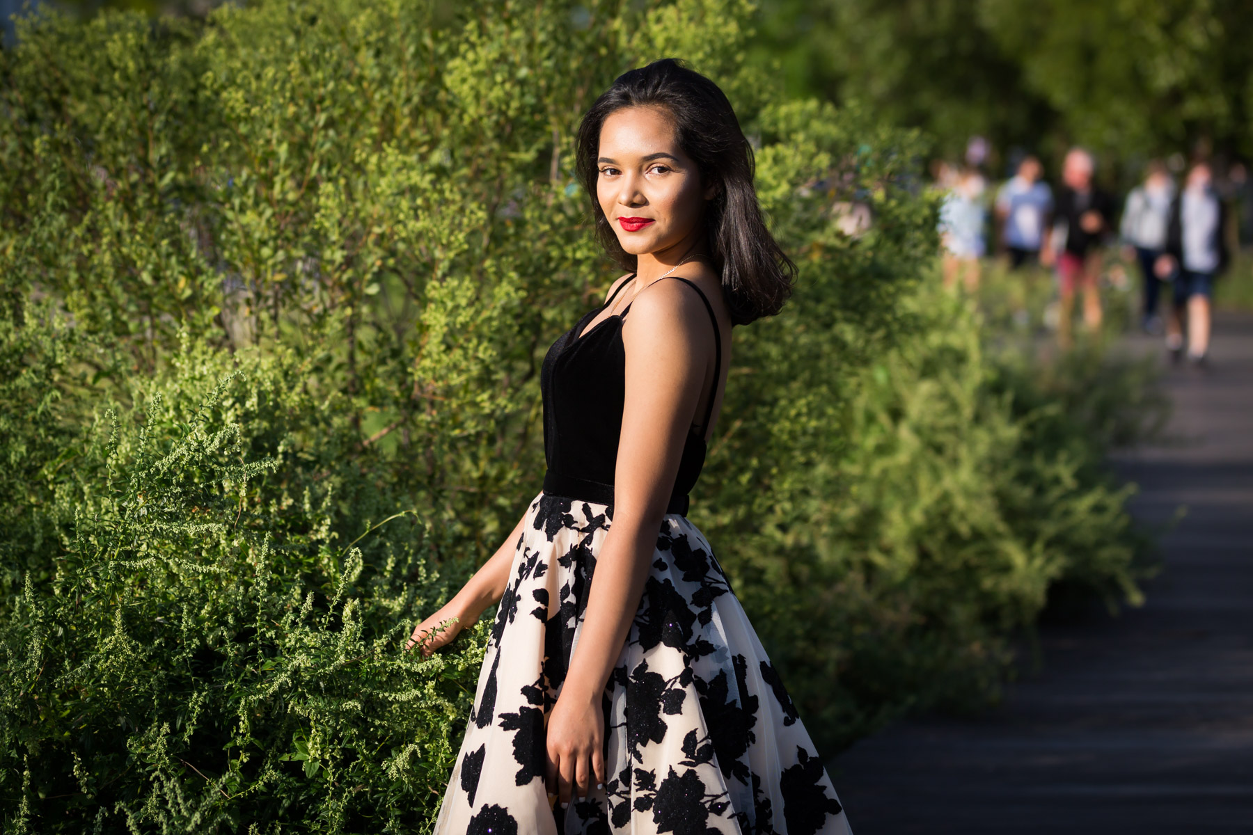 Smiling woman with brown hair and red lipstick standing in front of green bush for an article on how to beat the summer heat during a photo shoot