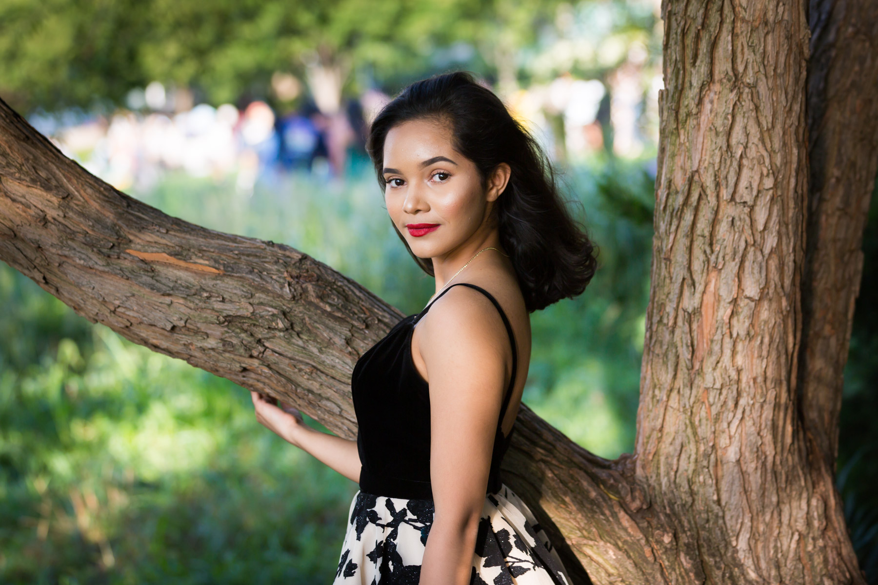 Smiling woman with brown hair and red lipstick leaning on tree for an article on how to beat the summer heat during a photo shoot