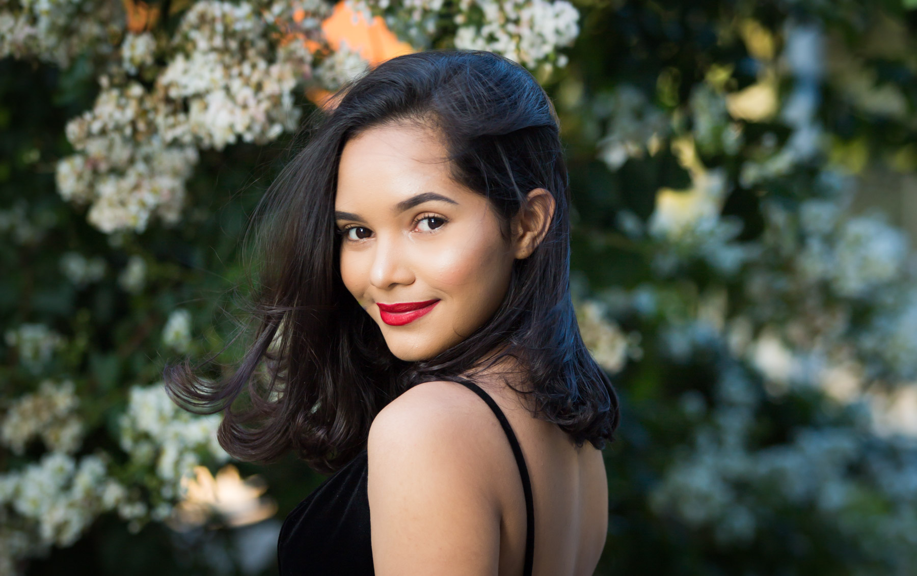 Smiling woman with brown hair and red lipstick looking over shoulder in front of bush for an article on how to beat the summer heat during a photo shoot