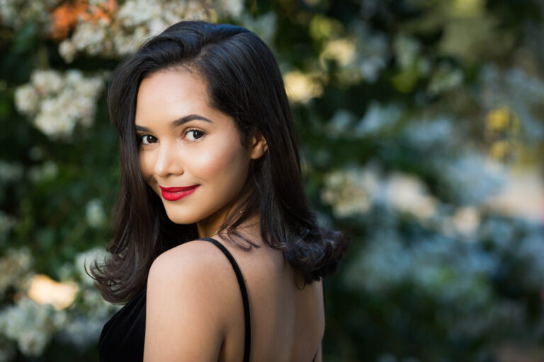 Smiling woman with brown hair and red lipstick looking over shoulder in front of bush for an article on how to beat the summer heat during a photo shoot