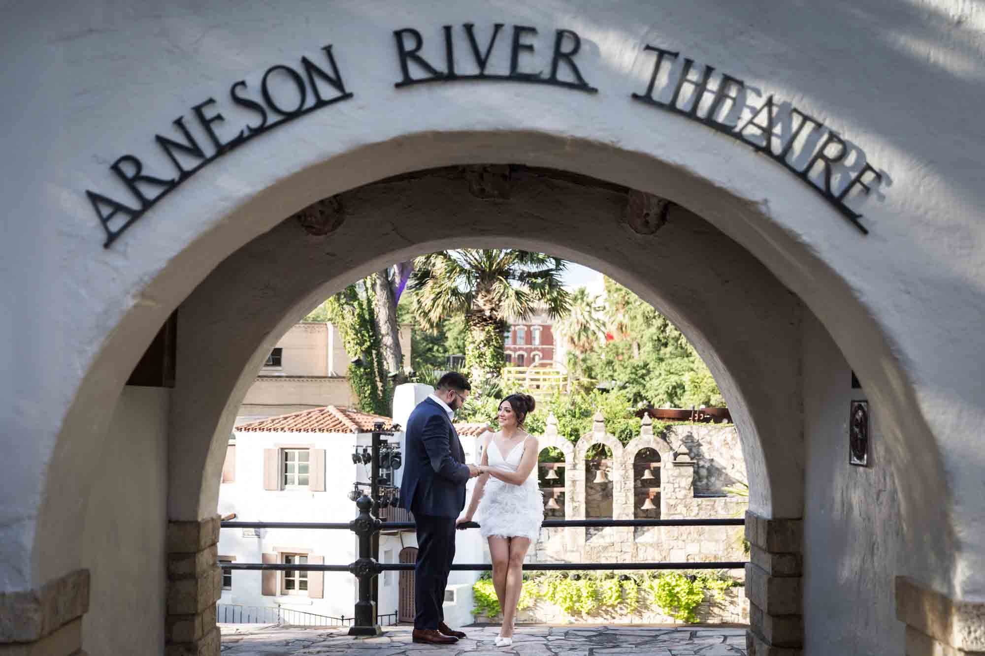 La Villita engagement photos of couple holding hands against railing shot through entrance to Arneson River Theatre