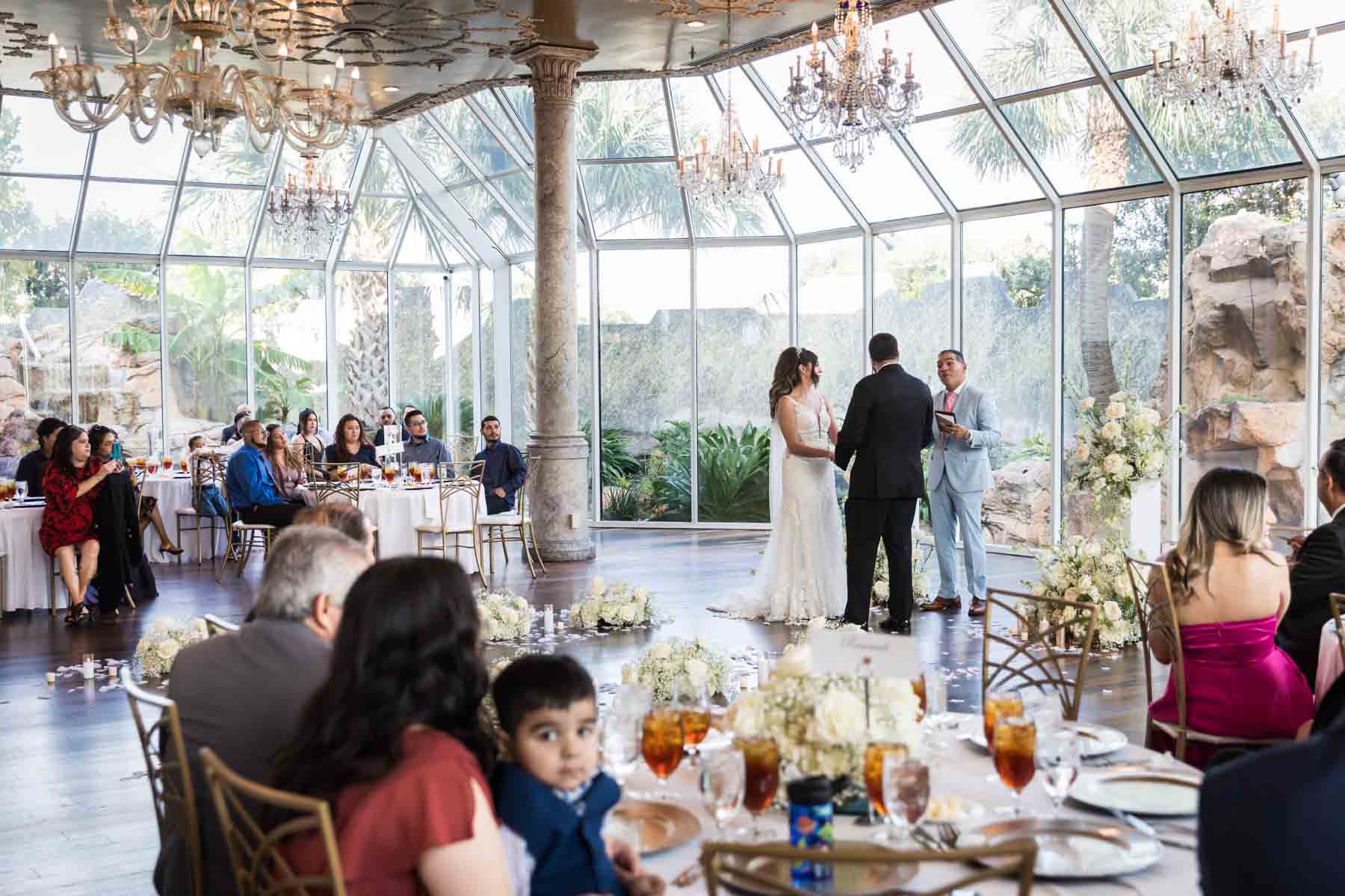 Old San Francisco wedding photos of bride and groom with officiant during ceremony surrounded by tables of guests in glass room