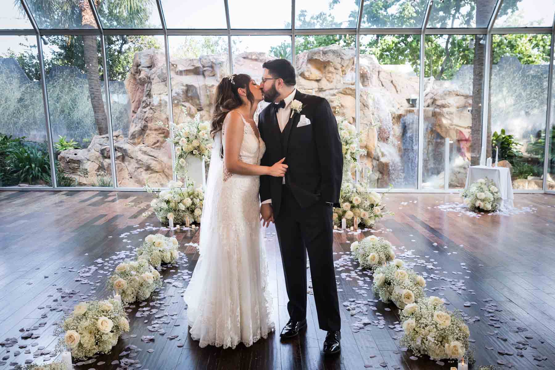 Old San Francisco wedding photos of bride and groom kissing ceremony surrounded by aisle of flowers in front of glass wall