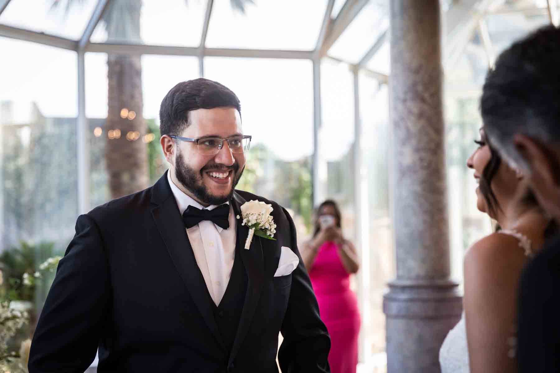 Groom wearing glasses and black tuxedo smiling at bride during ceremony at an Old San Francisco Steakhouse wedding