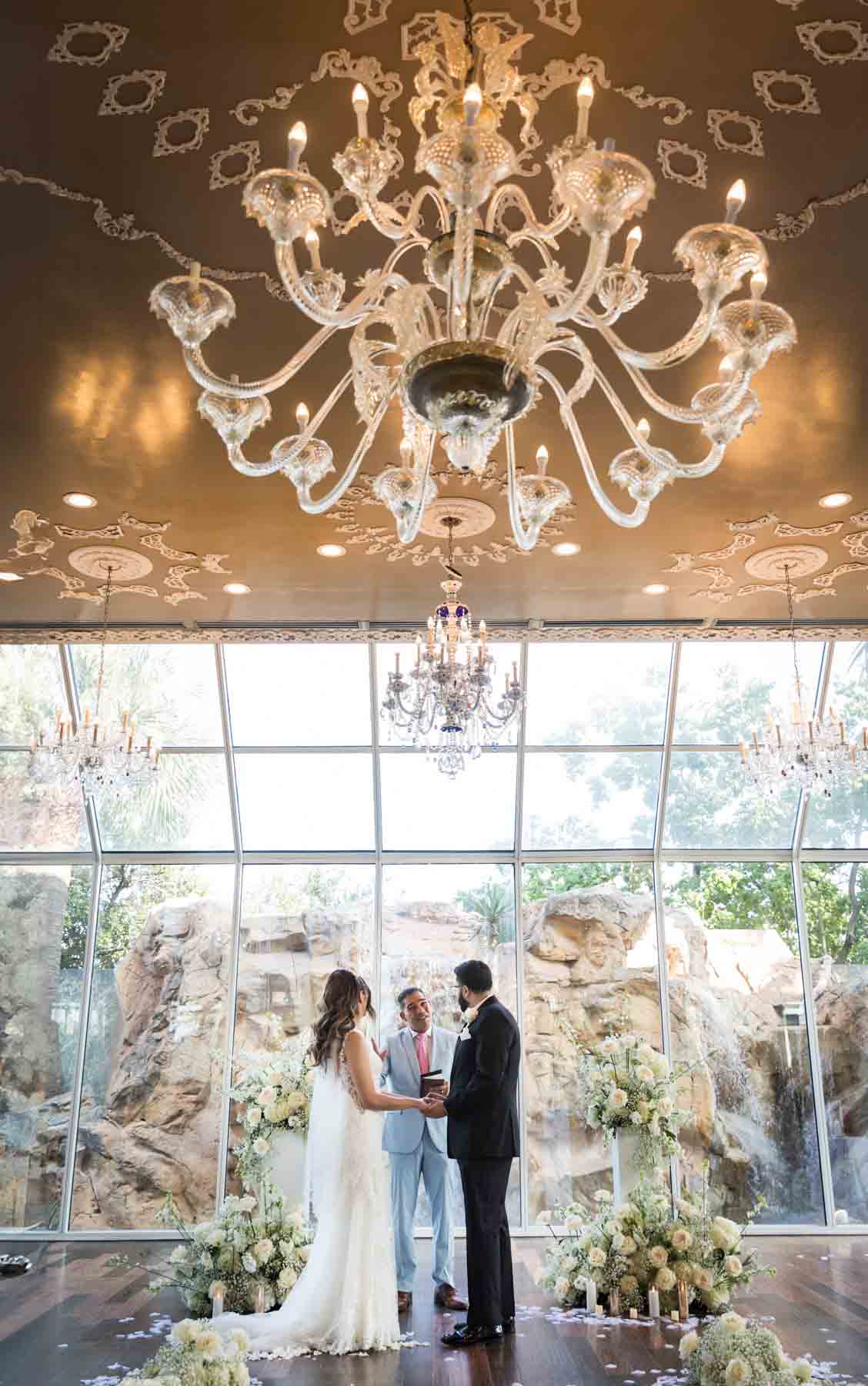 Old San Francisco wedding photos of bride and groom holding hands during ceremony with large chandelier overhead