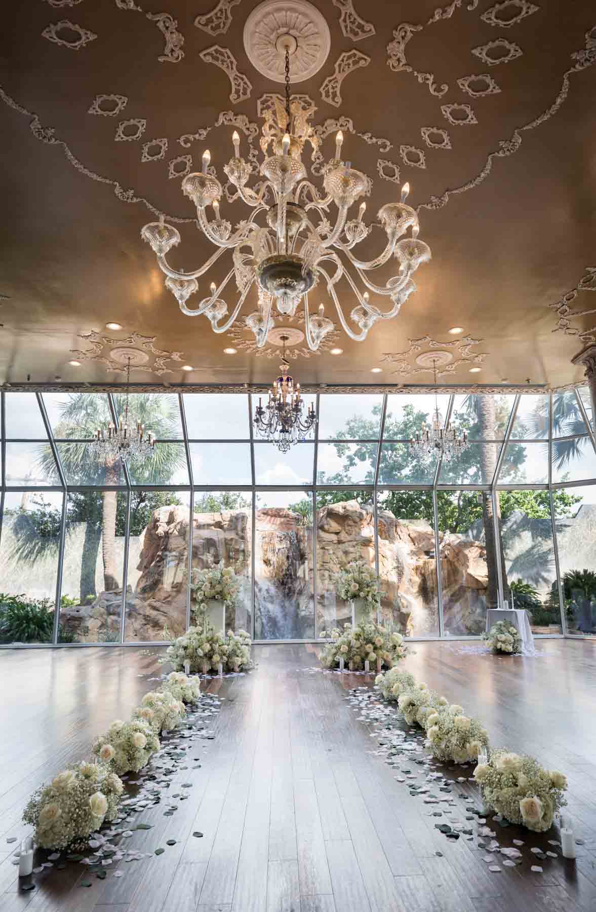 Aisle on wood floor with white flowers in front of glass wall and chandelier overhead at an Old San Francisco Steakhouse wedding