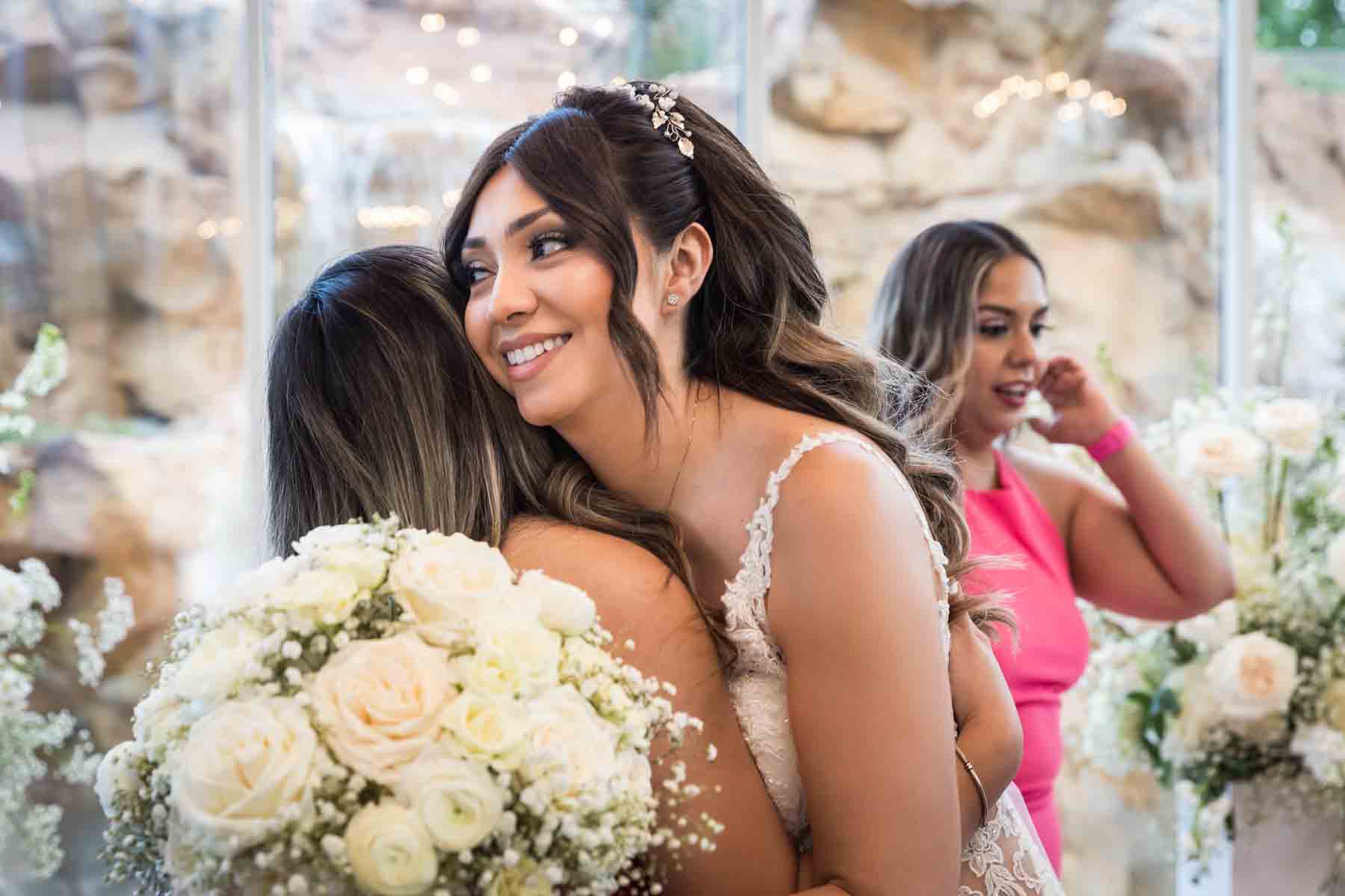 Bride hugging female guest with white bouquet in front and glass wall in back at an Old San Francisco Steakhouse wedding