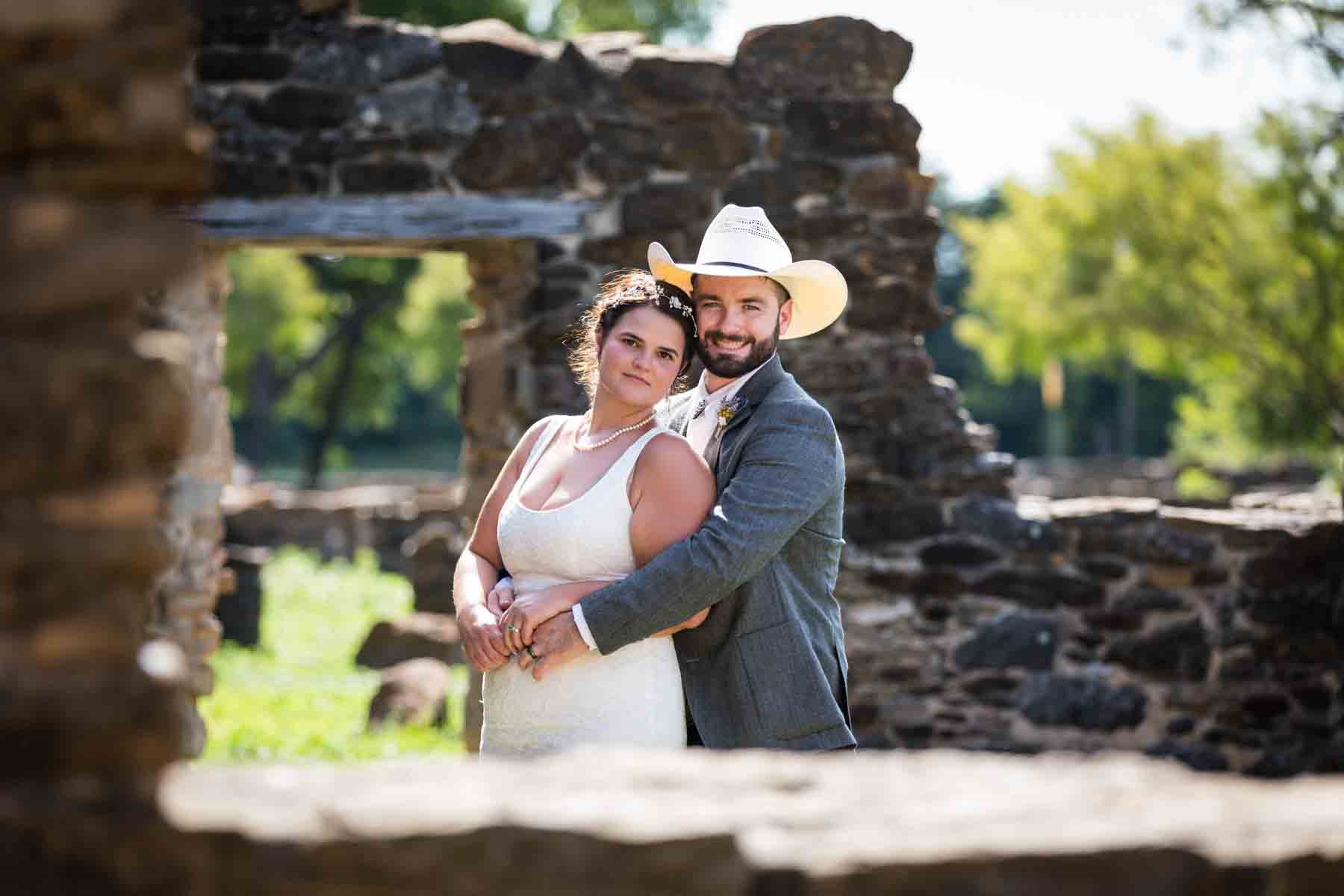 Bride and groom wearing cowboy hat hugging in front of stone ruins of Mission Espada