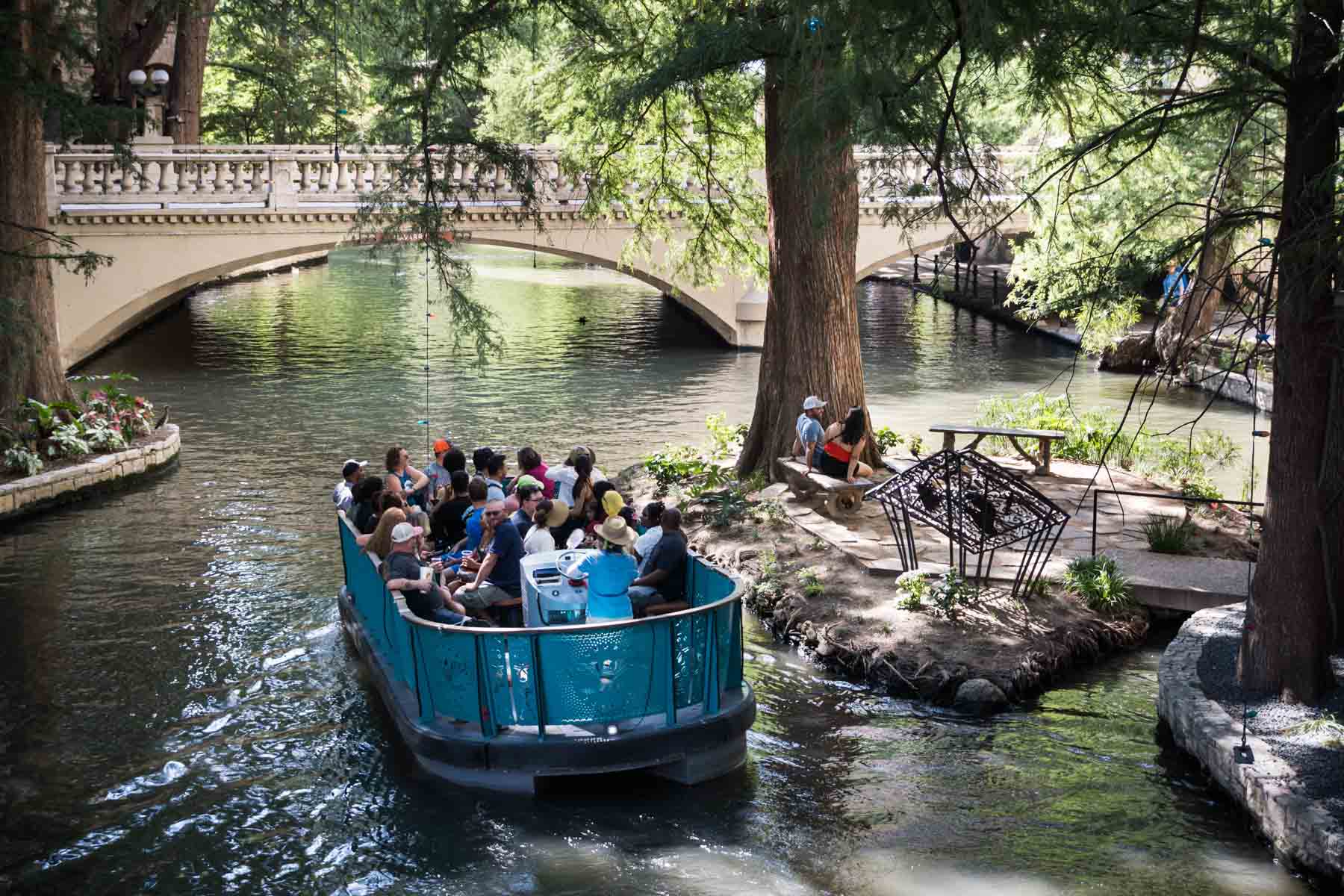 Couple sitting on bench on Marriage Island with blue River Walk shuttle floating past for an article listing the best places to propose in San Antonio