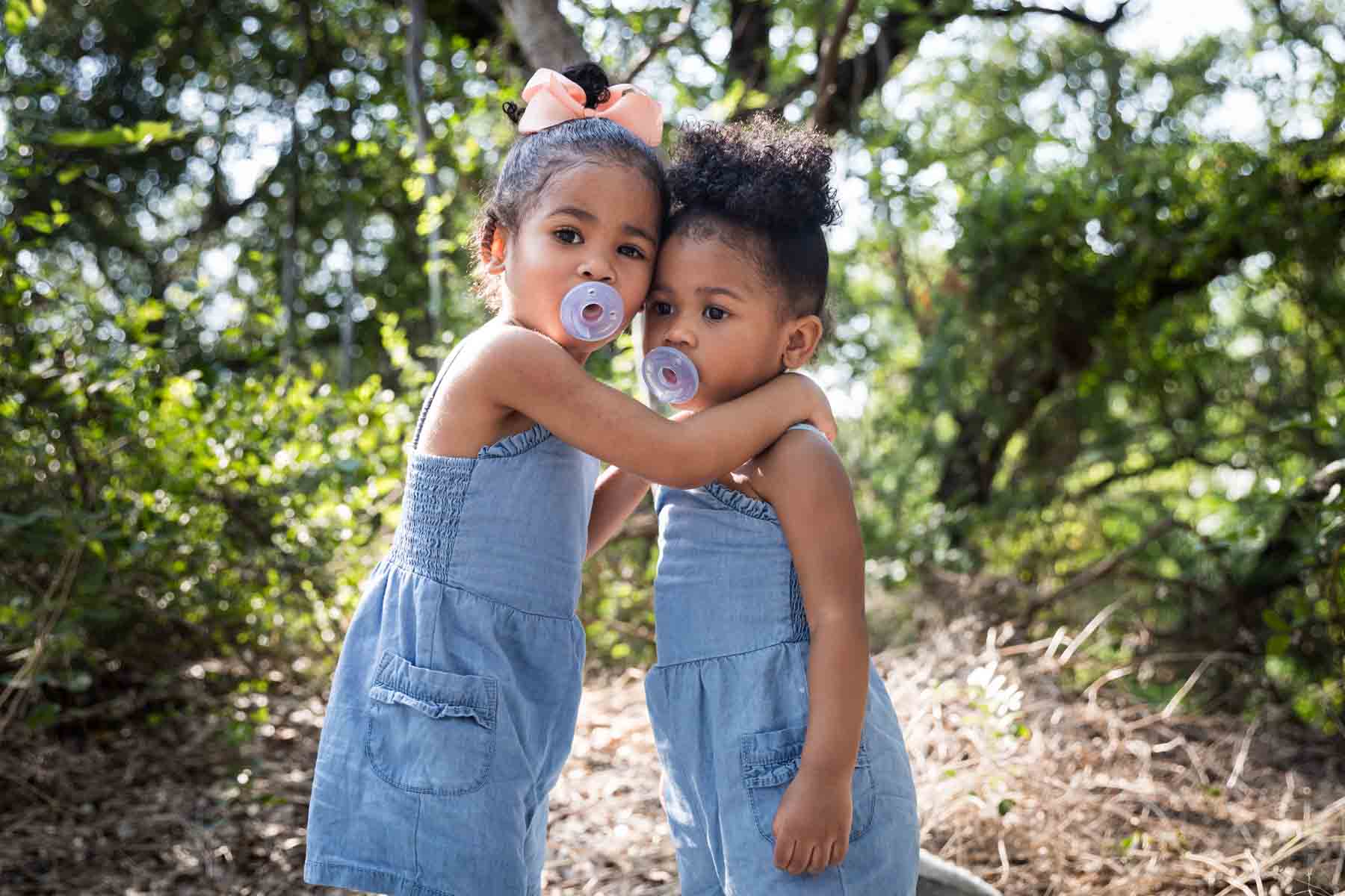 Two little girls sucking on pacifiers and hugging each other in forest of Brackenridge Park