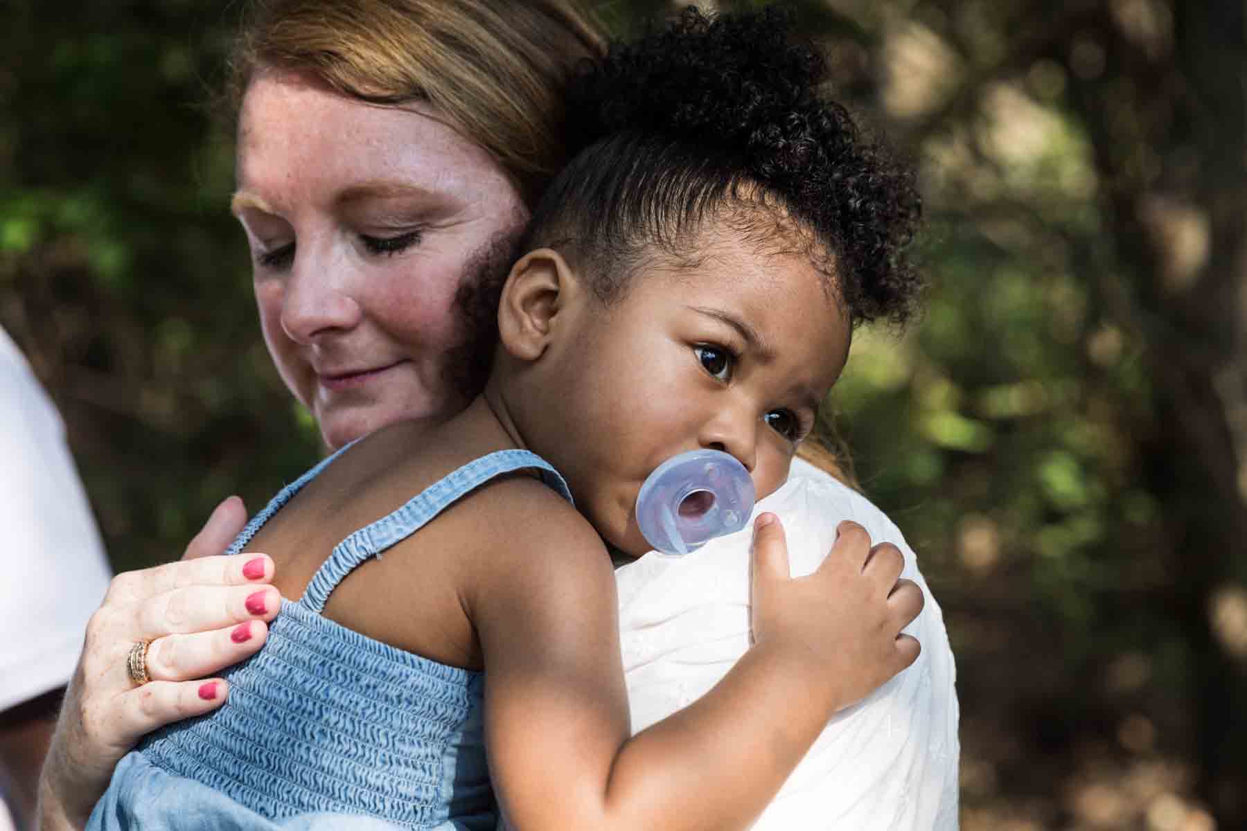 Grandmother holding adorable little girl on her shoulder sucking on a pacifier in a forest