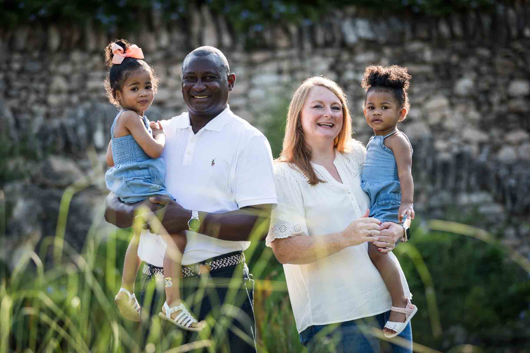 Mixed race couple holding two adorable little girls in front of stone wall at the Japanese Tea Garden for an article on 'How often should I take family photos?'