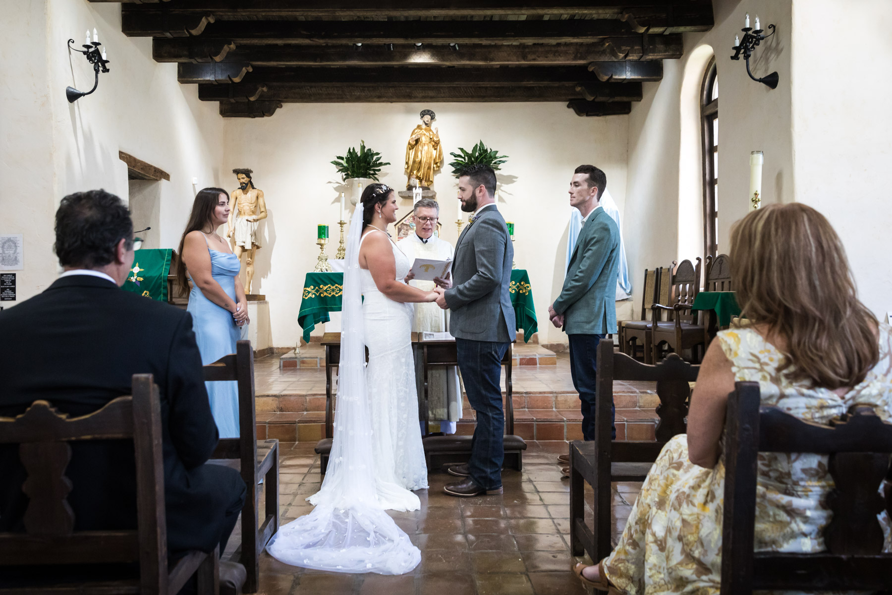 Bride and groom holding hands in front of altar and bridal party for an article on how to get married at Mission Espada