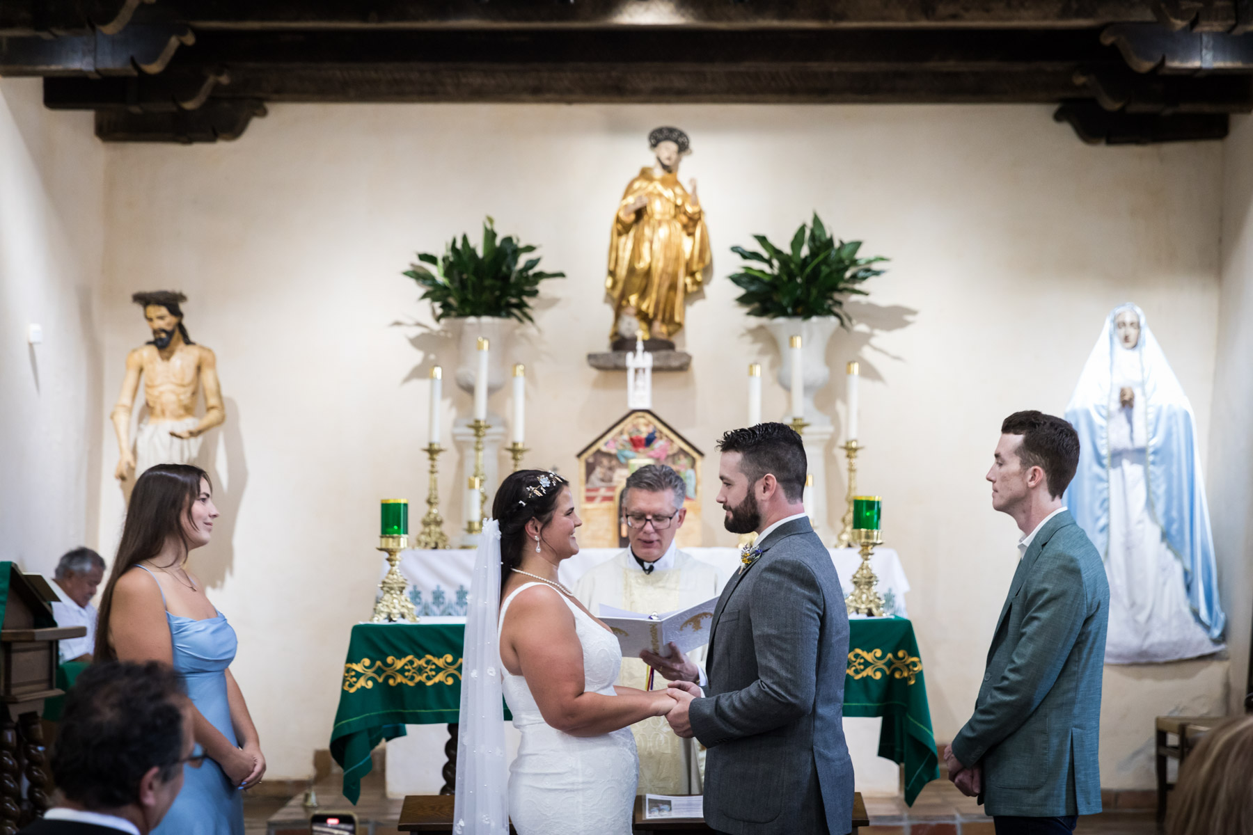 Bride and groom holding hands in front of bridal party and priest during ceremony for an article on how to get married at Mission Espada