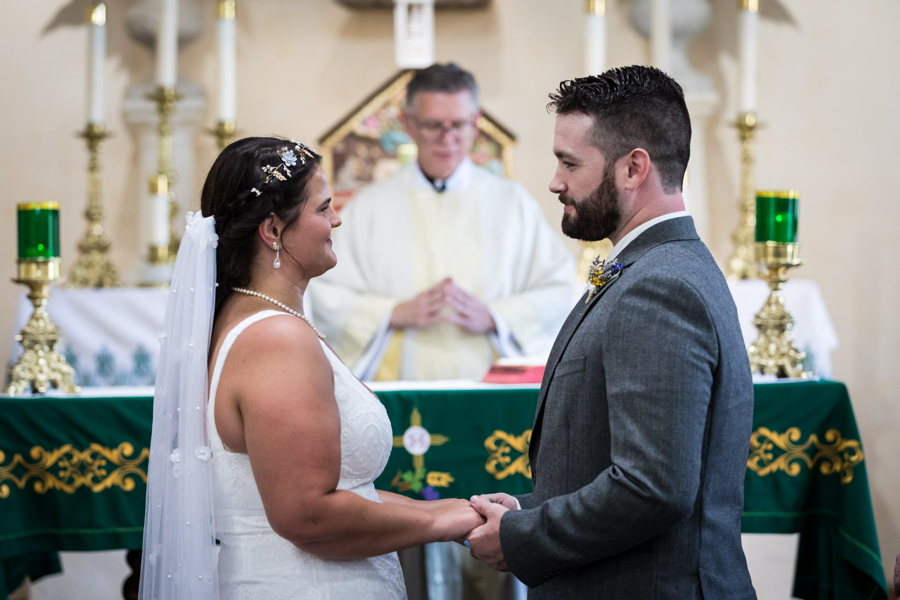 Bride and groom holding hands and smiling at each other in front of altar and priest for an article on how to get married at Mission Espada