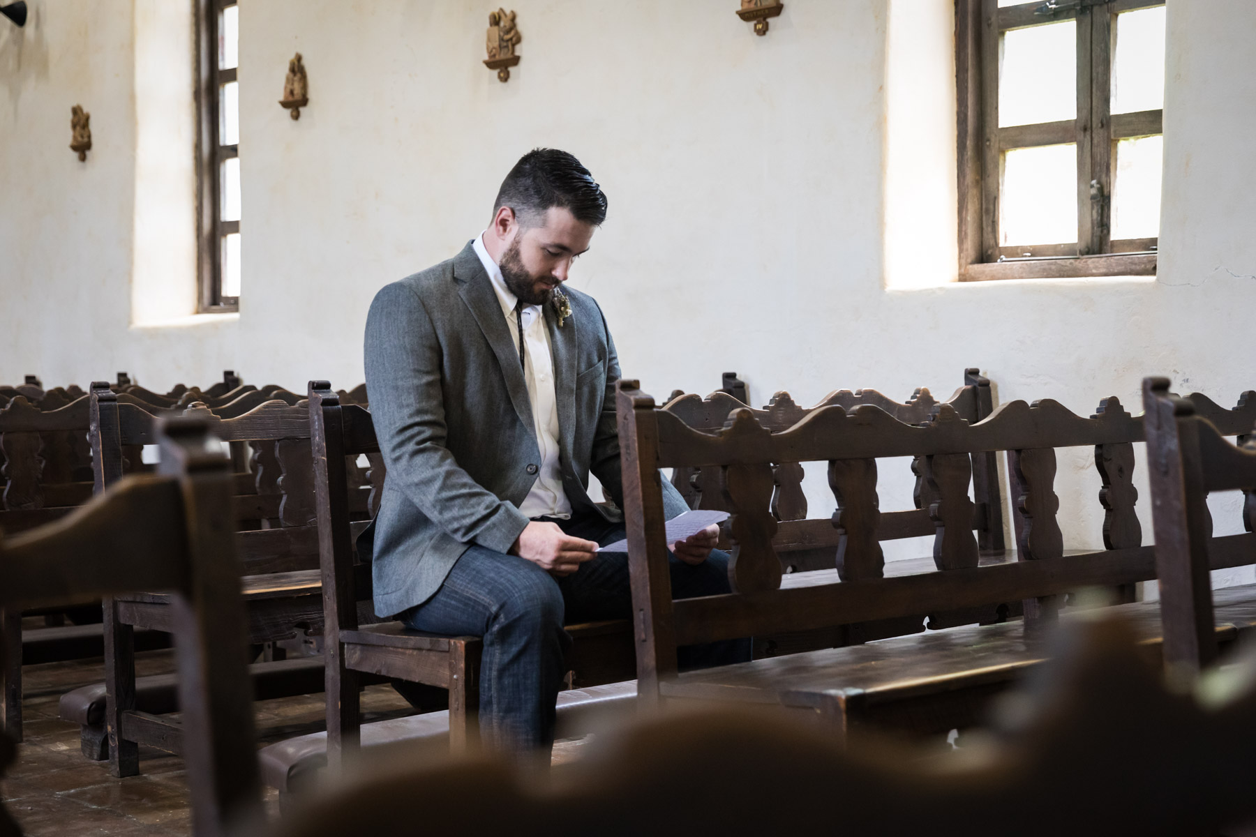 Groom wearing grey jacket sitting in wooden pew reading letter