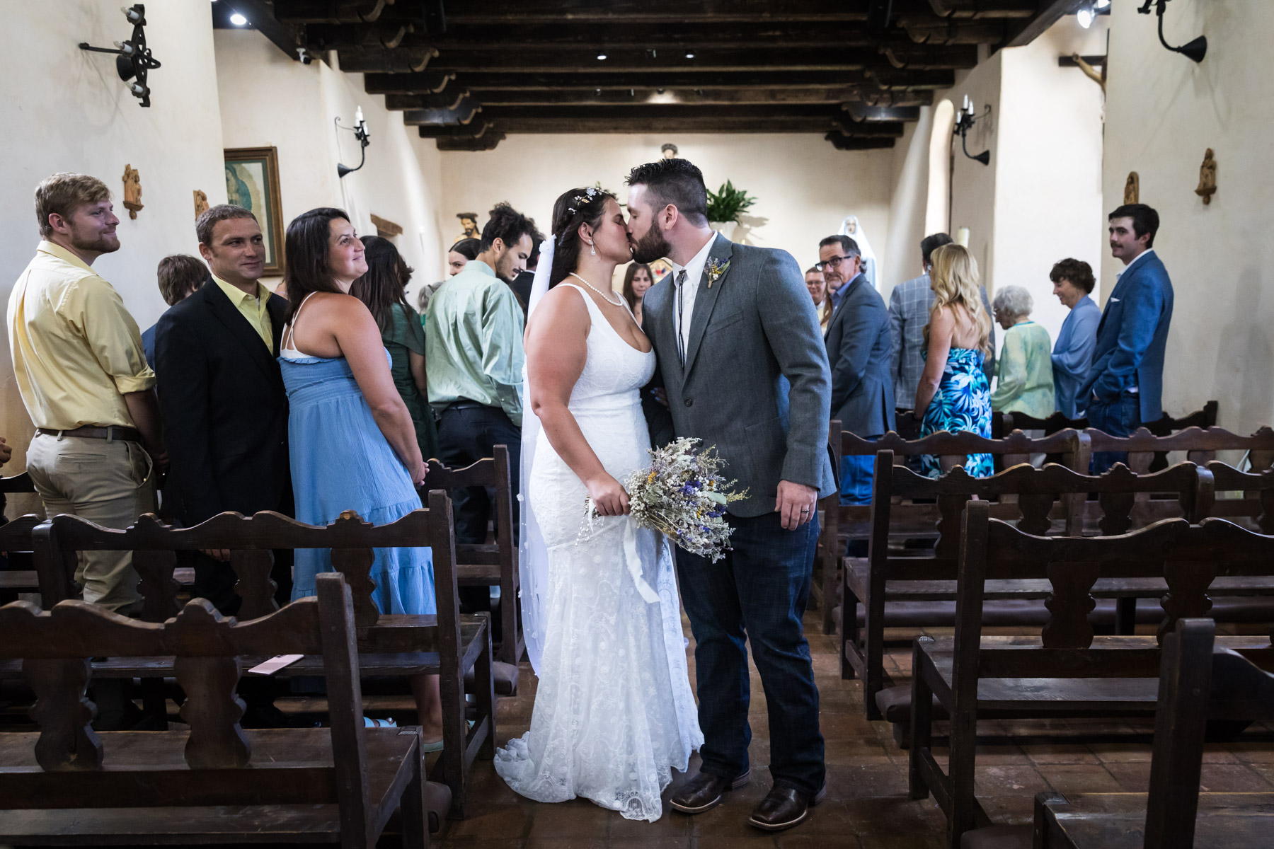 Bride and groom kissing in aisle surrounded by guests for an article on how to get married at Mission Espada