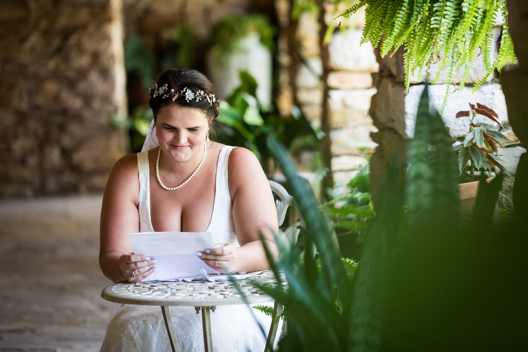 Bride wearing white sleeveless dress sitting at outside table reading letter