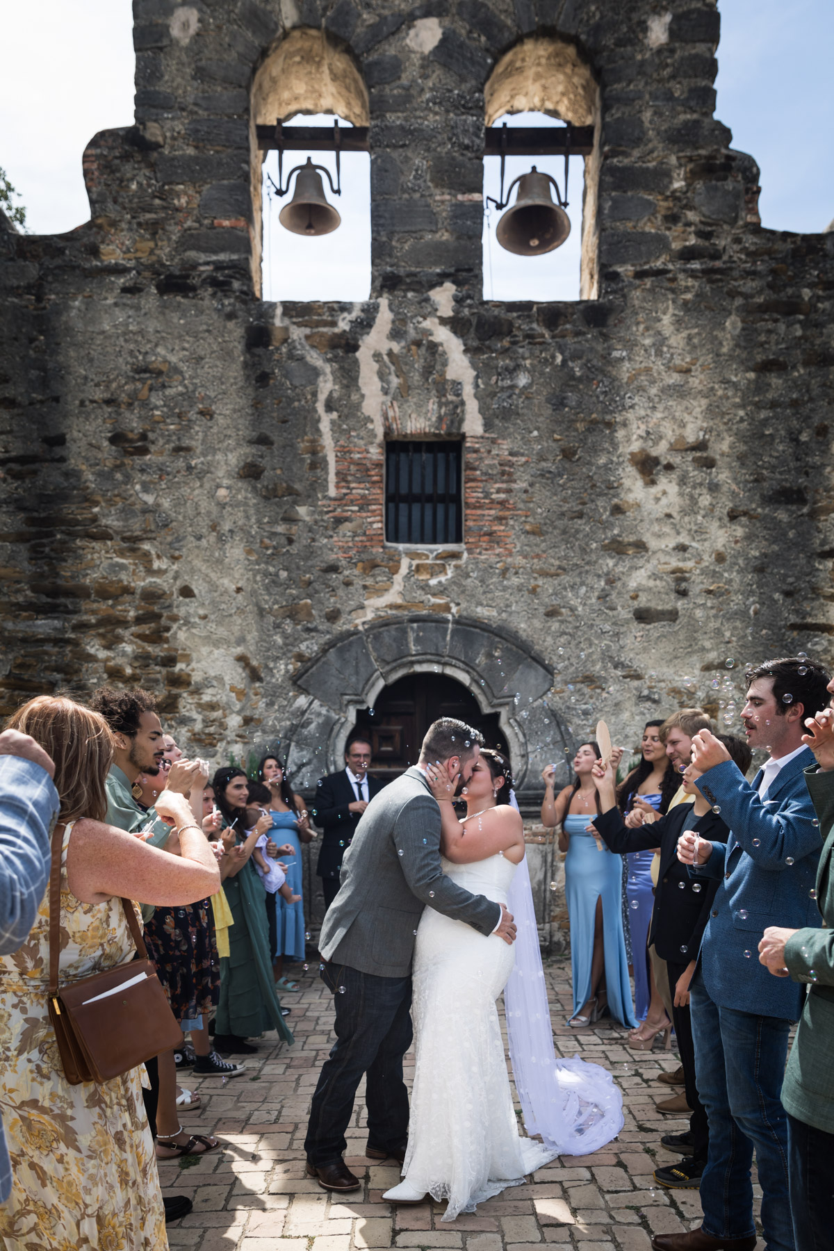 Bride and groom kissing in row of guests blowing bubbles for an article on how to get married at Mission Espada