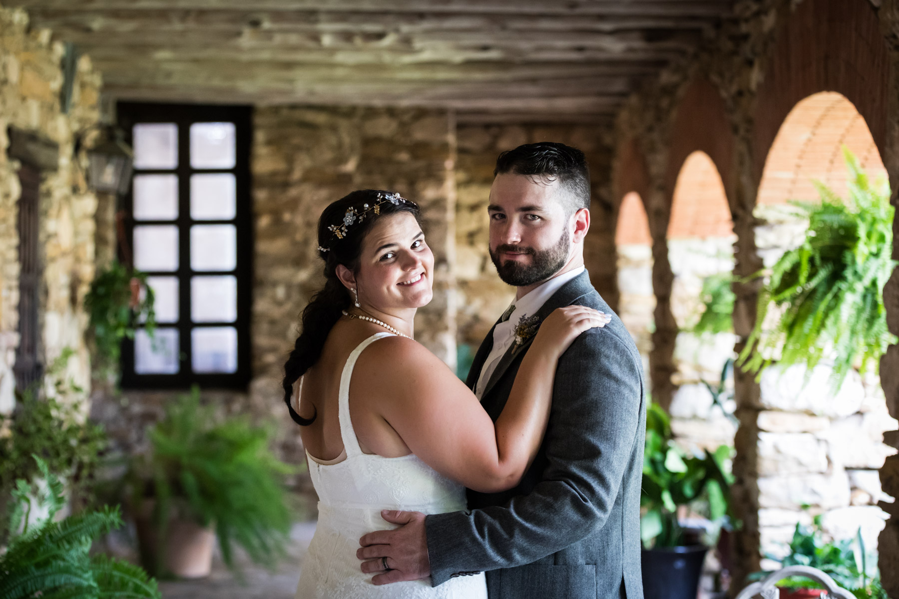 Bride in white sleeveless dress and groom wearing grey jacket hugging under covered walkway for an article on how to get married at Mission Espada