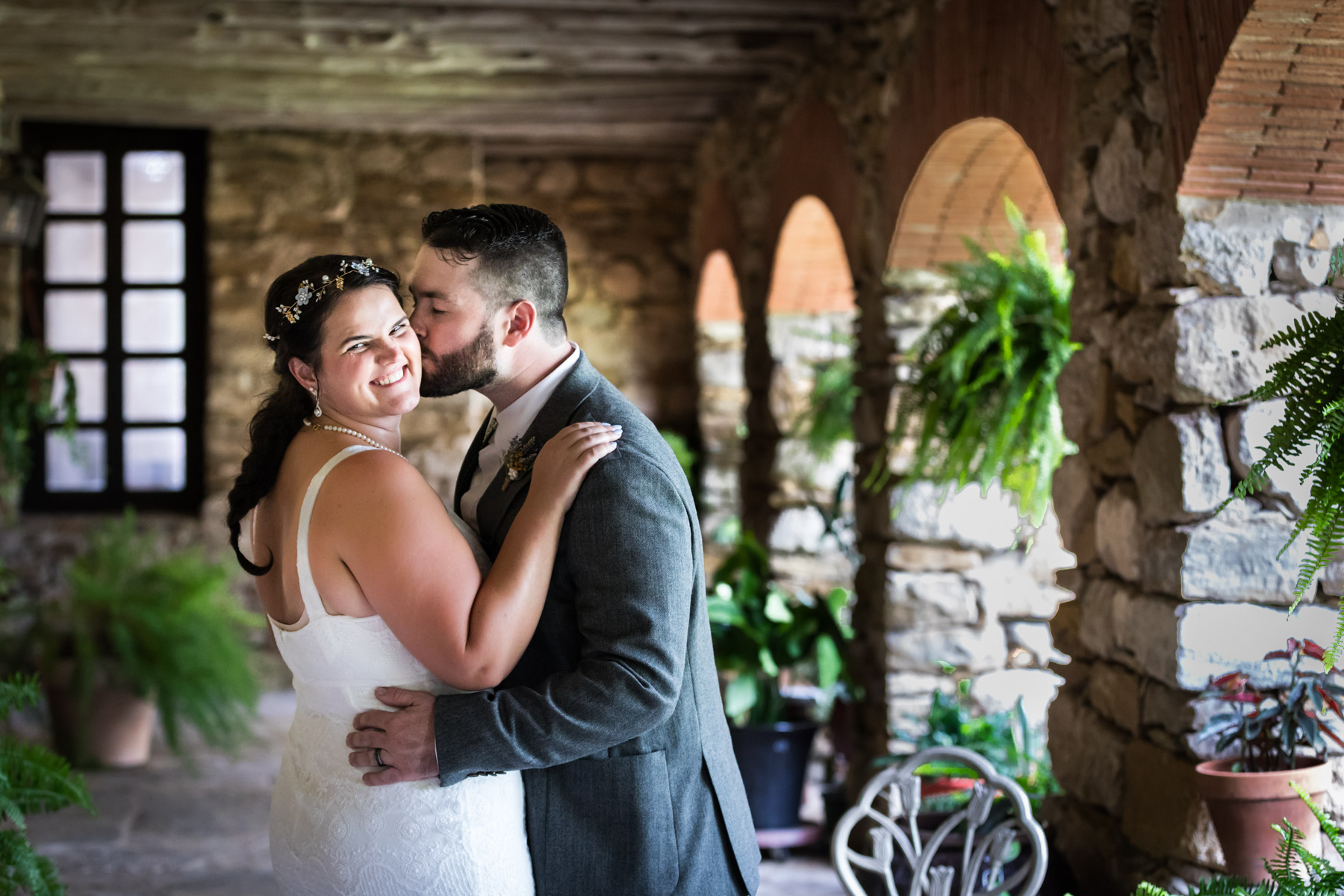 Bride in white sleeveless dress and groom wearing grey jacket kissing under covered walkway for an article on how to get married at Mission Espada