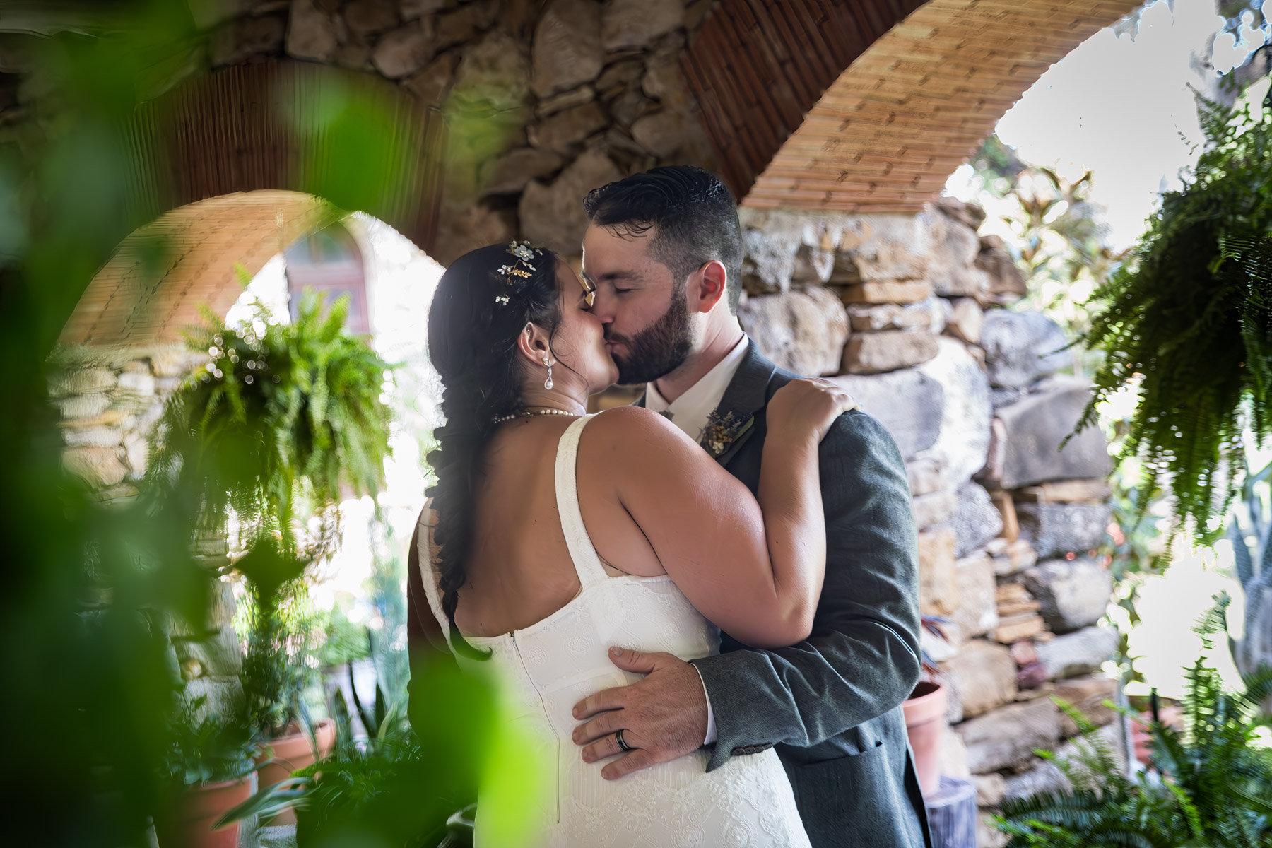 Bride in white sleeveless dress and groom wearing grey jacket kissing under covered walkway for an article on how to get married at Mission Espada