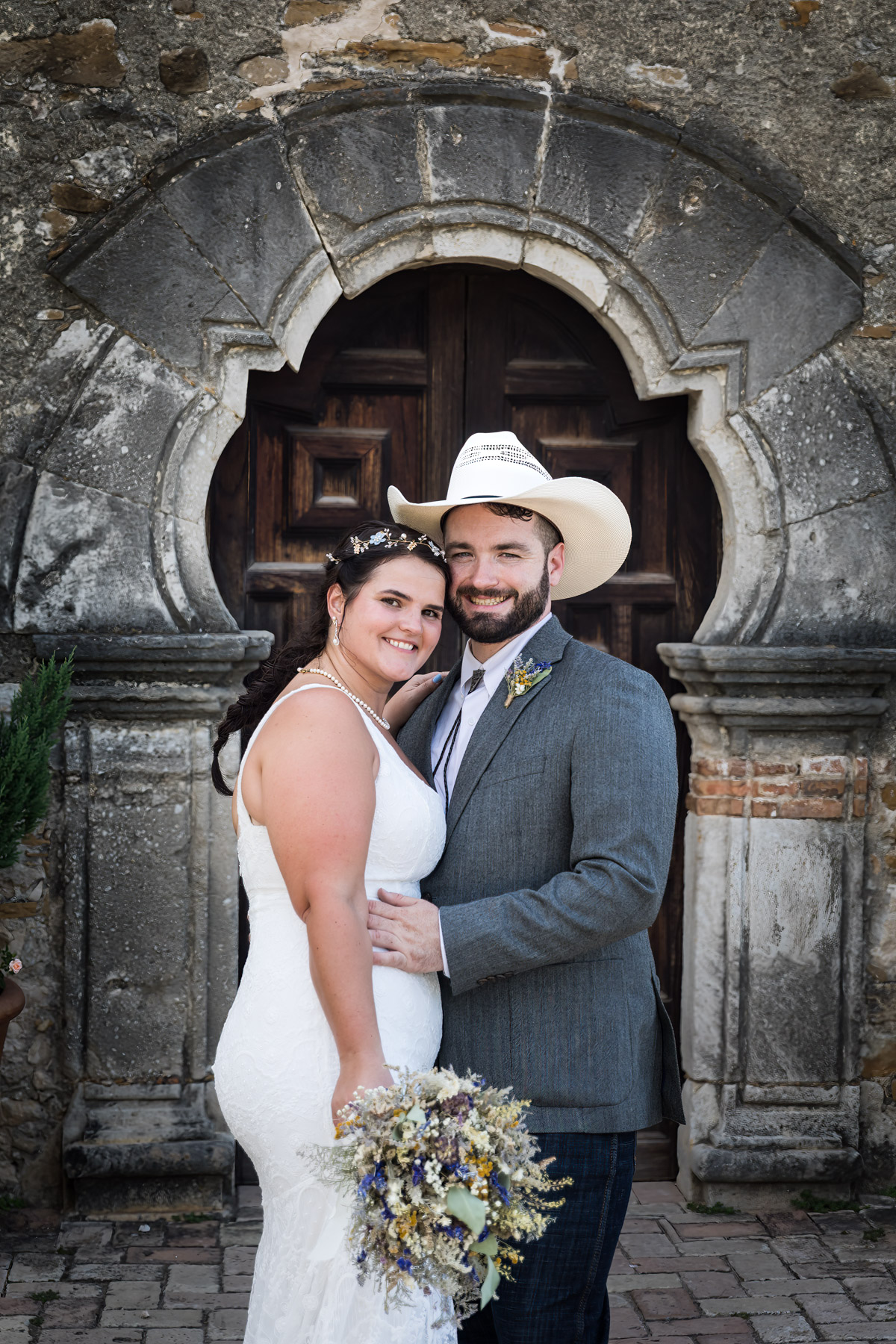 Bride in white sleeveless dress and groom wearing cowboy hat in front of ornate wooden door for an article on how to get married at Mission Espada