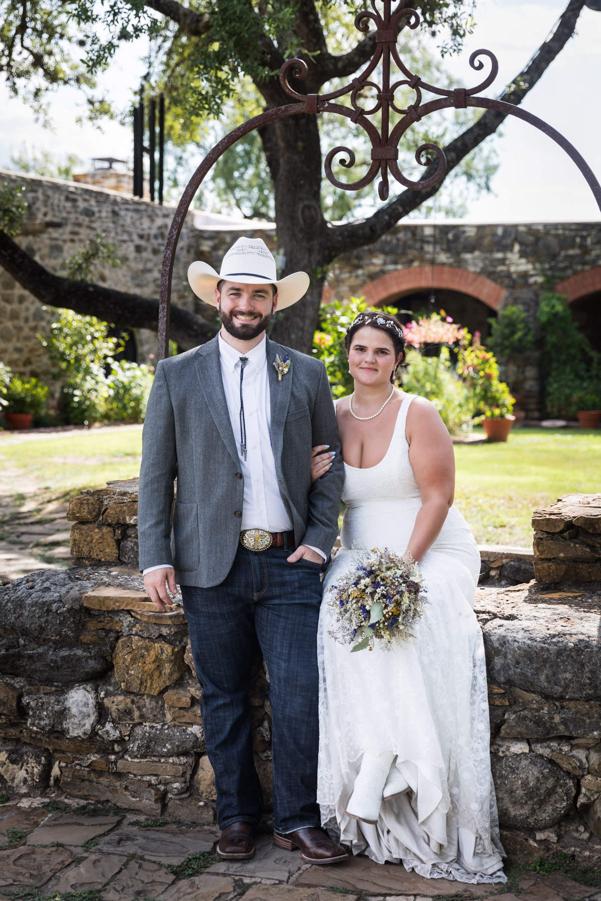 Bride in white sleeveless dress and groom wearing cowboy hat in front of well for an article on how to get married at Mission Espada