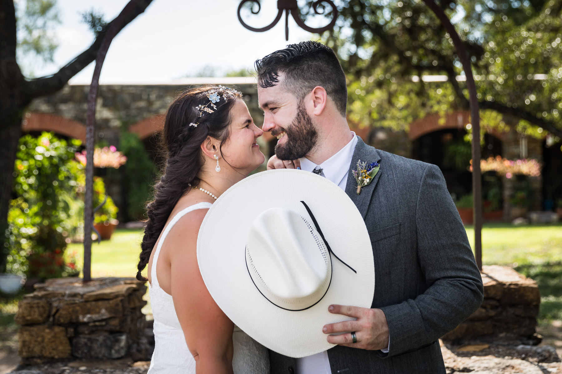 Bride and groom smiling at each other behind cowboy hat for an article on how to get married at Mission Espada