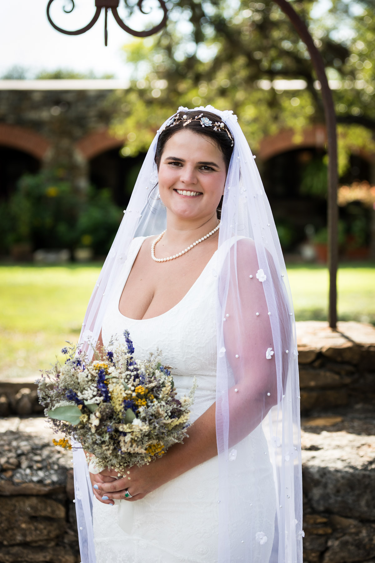 Bride wearing white sleeveless dress and veil holding flower bouquet at Mission Espada