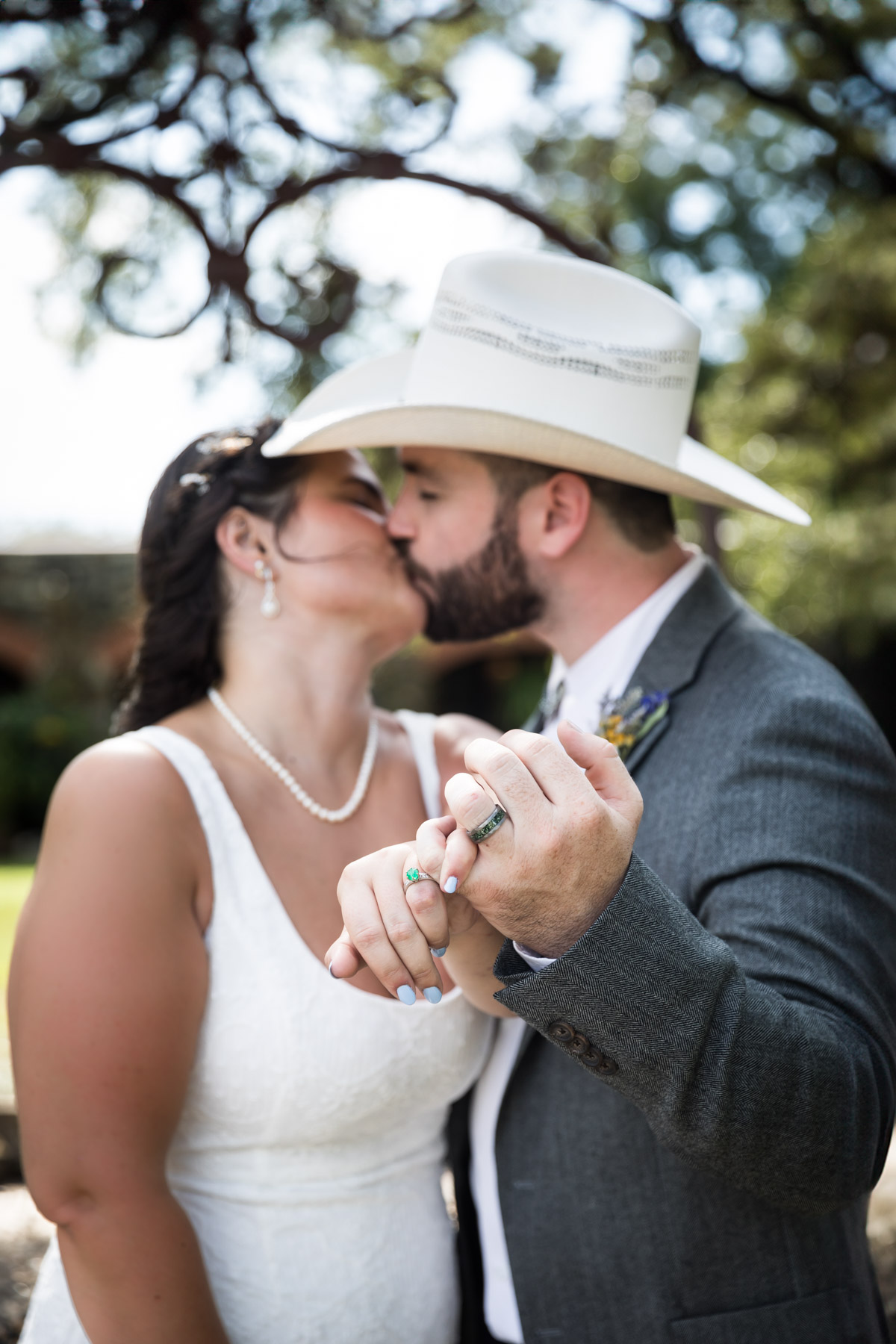 Bride in white sleeveless dress and groom wearing grey jacket and cowboy hat kissing and holding out hands showing wedding rings