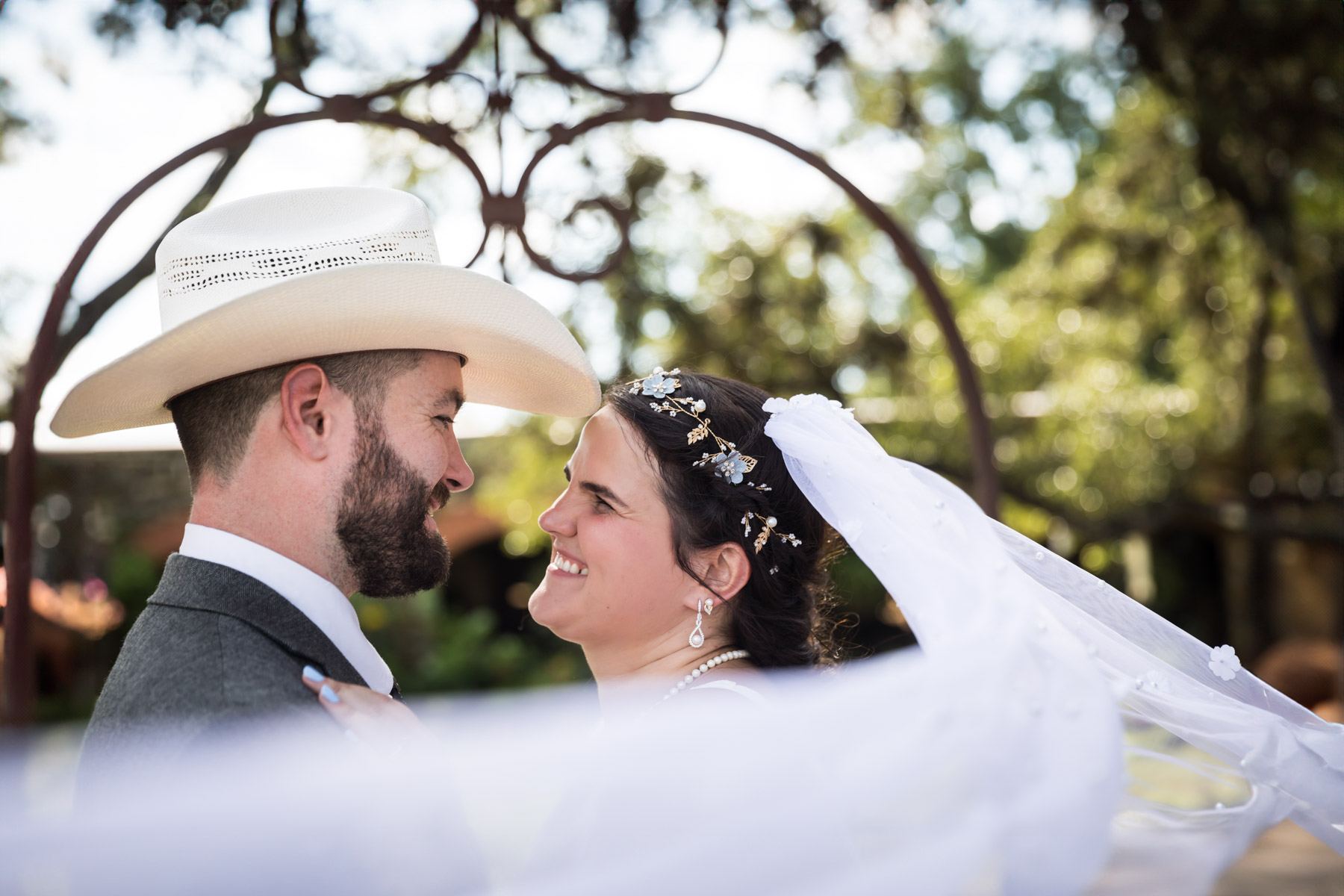 Bride in veil and groom wearing grey jacket and cowboy hat smiling at each other