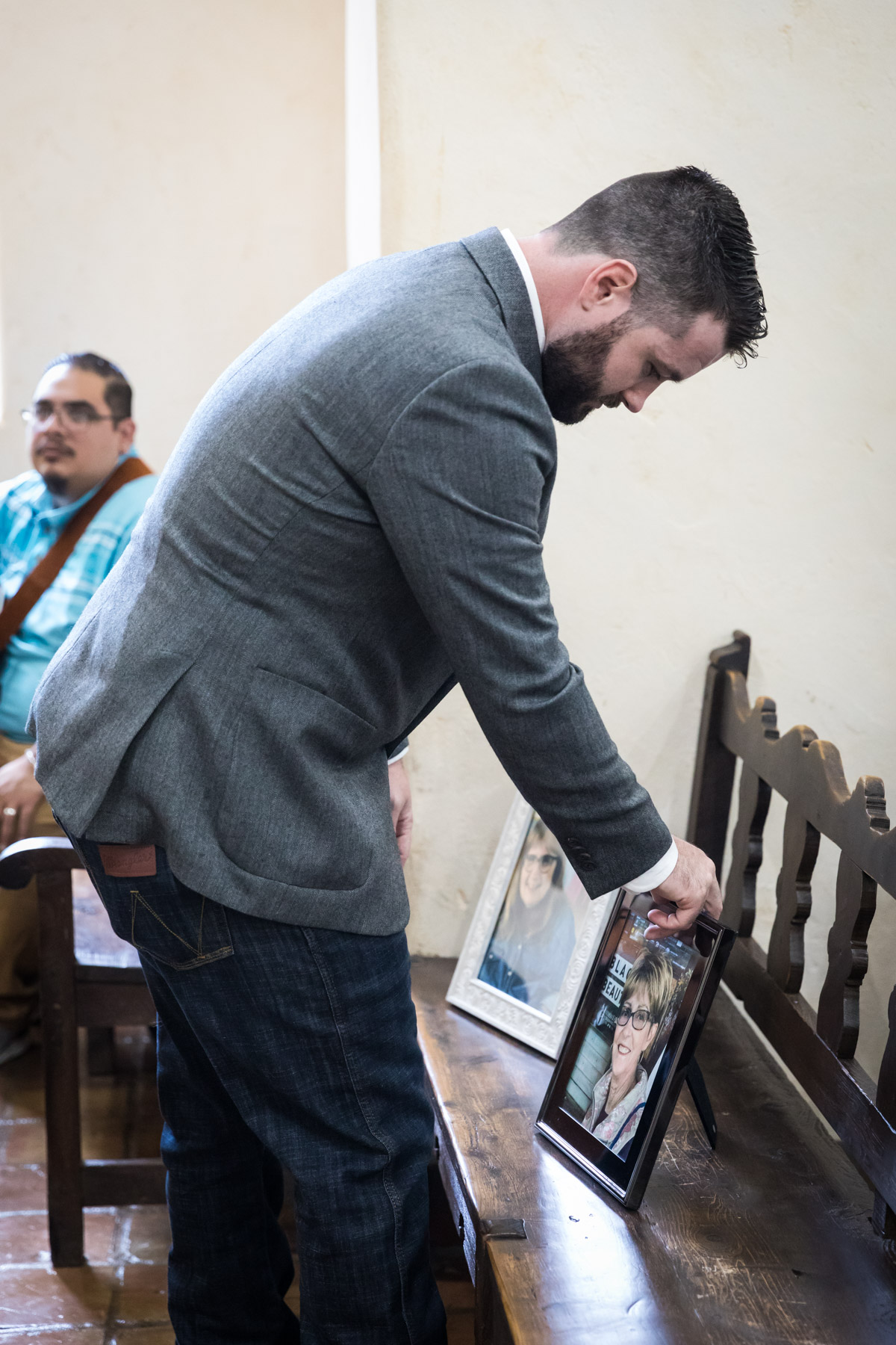 Groom adjusting framed portraits of two departed family members on wooden pew for an article on how to get married at Mission Espada