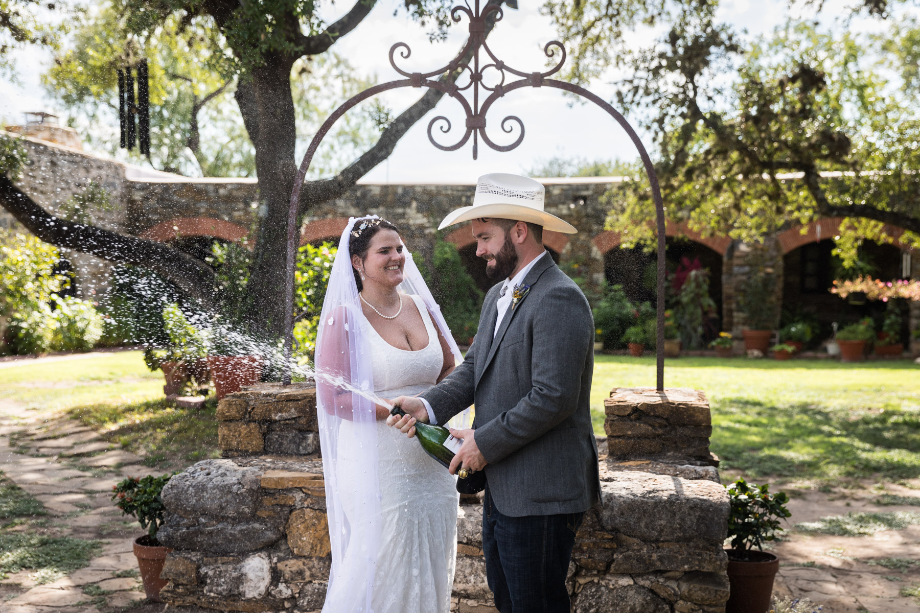 Bride in veil and groom wearing grey jacket and cowboy hat spraying bottle of champagne